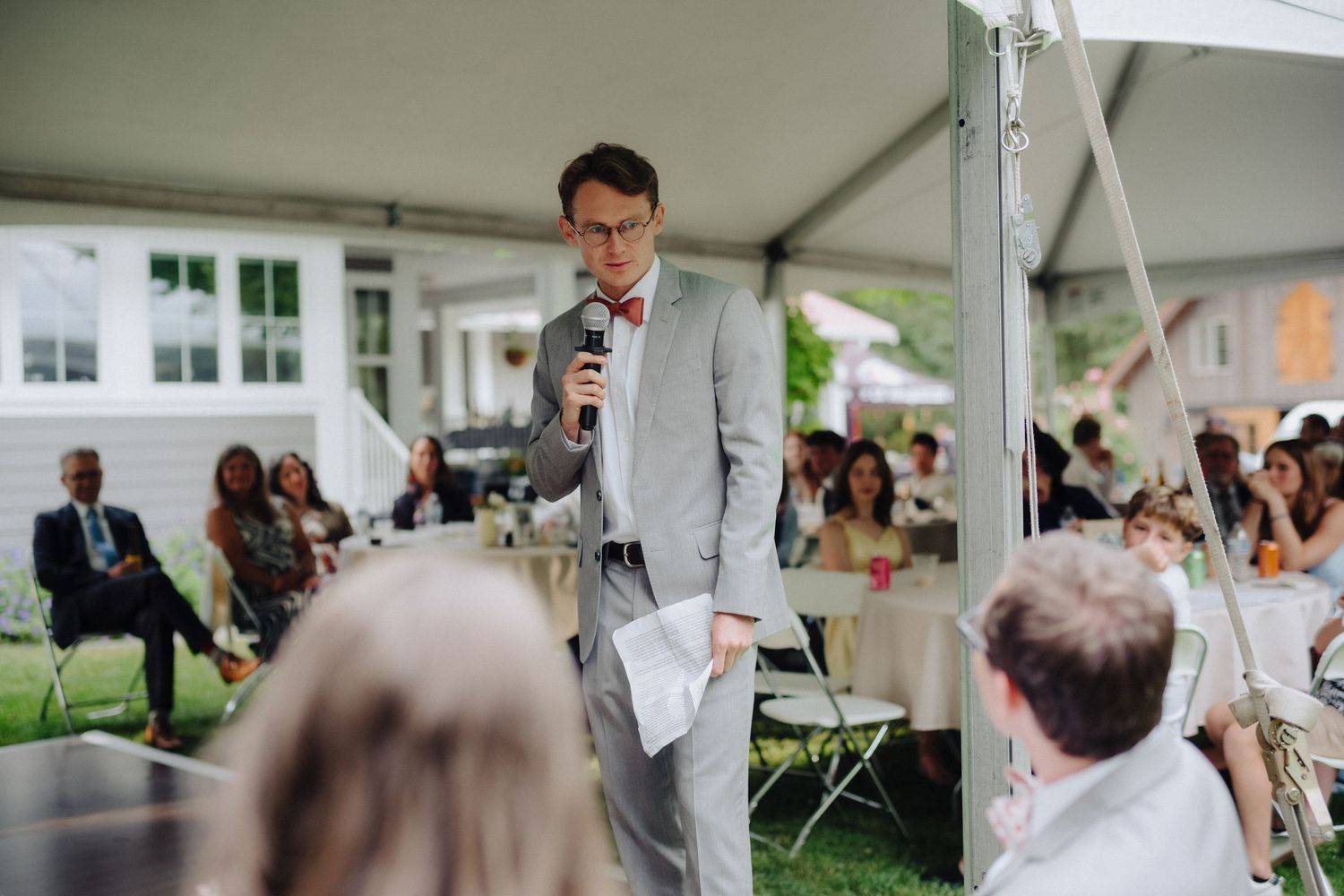 Wedding reception guests seated at round tables under white tent.