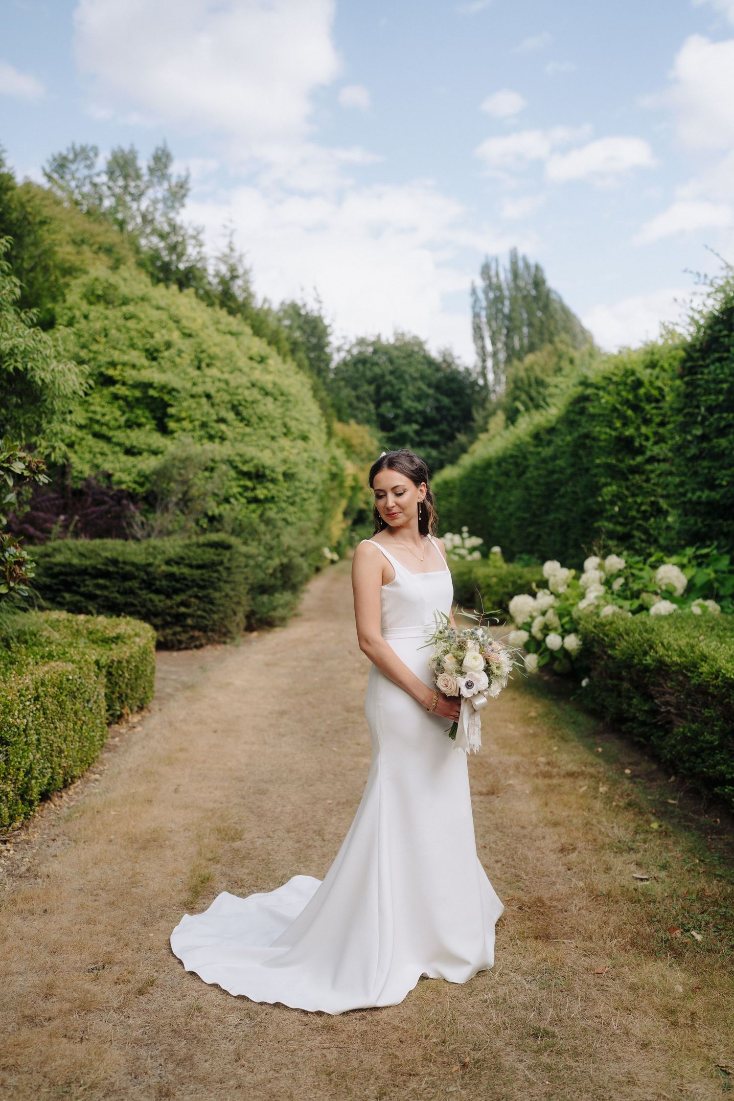 Elegant bride in white dress poses with bouquet along garden pathway surrounded by greenery.
