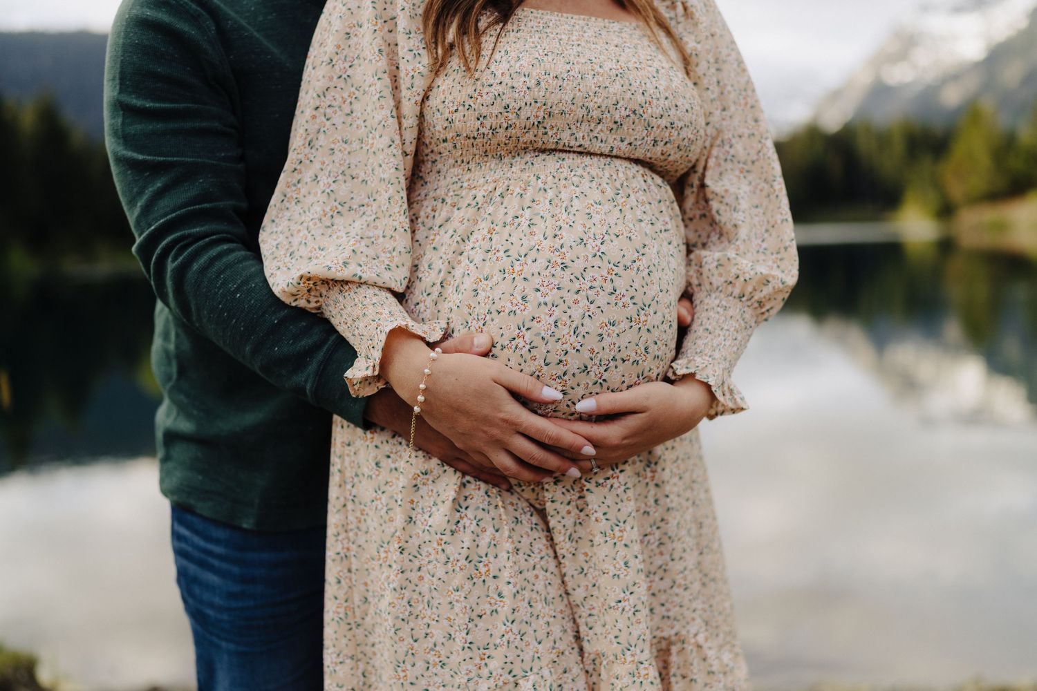 Intimate maternity photoshoot by a tranquil lake with mountains and forest creating a scenic backdrop.