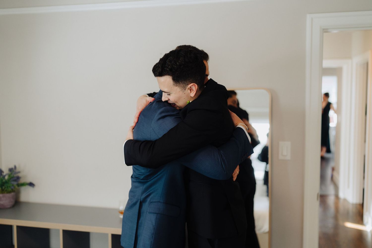 Two people share an emotional embrace in a bright, minimalist interior space while a third figure stands in the doorway.