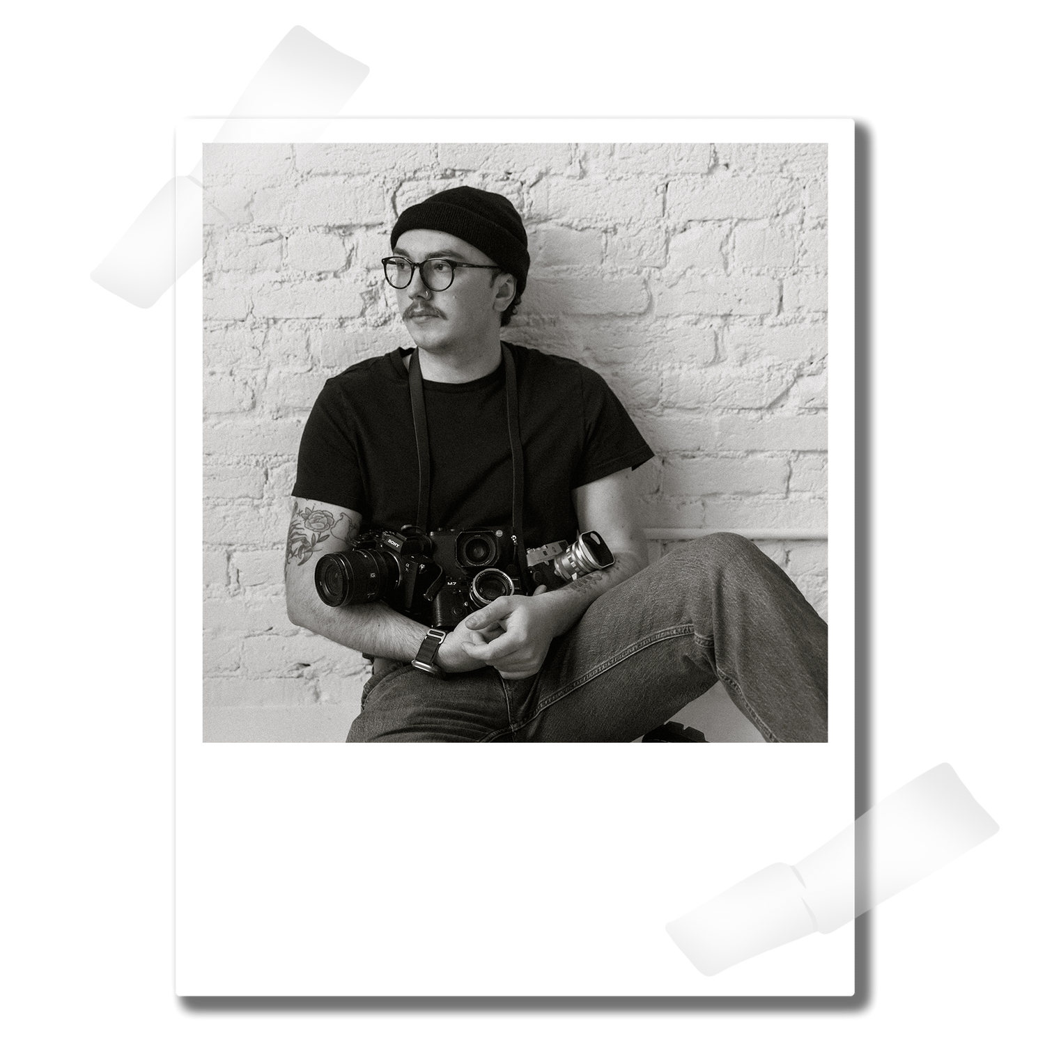 Black and white photo of a tattooed man sitting cross-legged holding a camera, wearing glasses and a black cap.