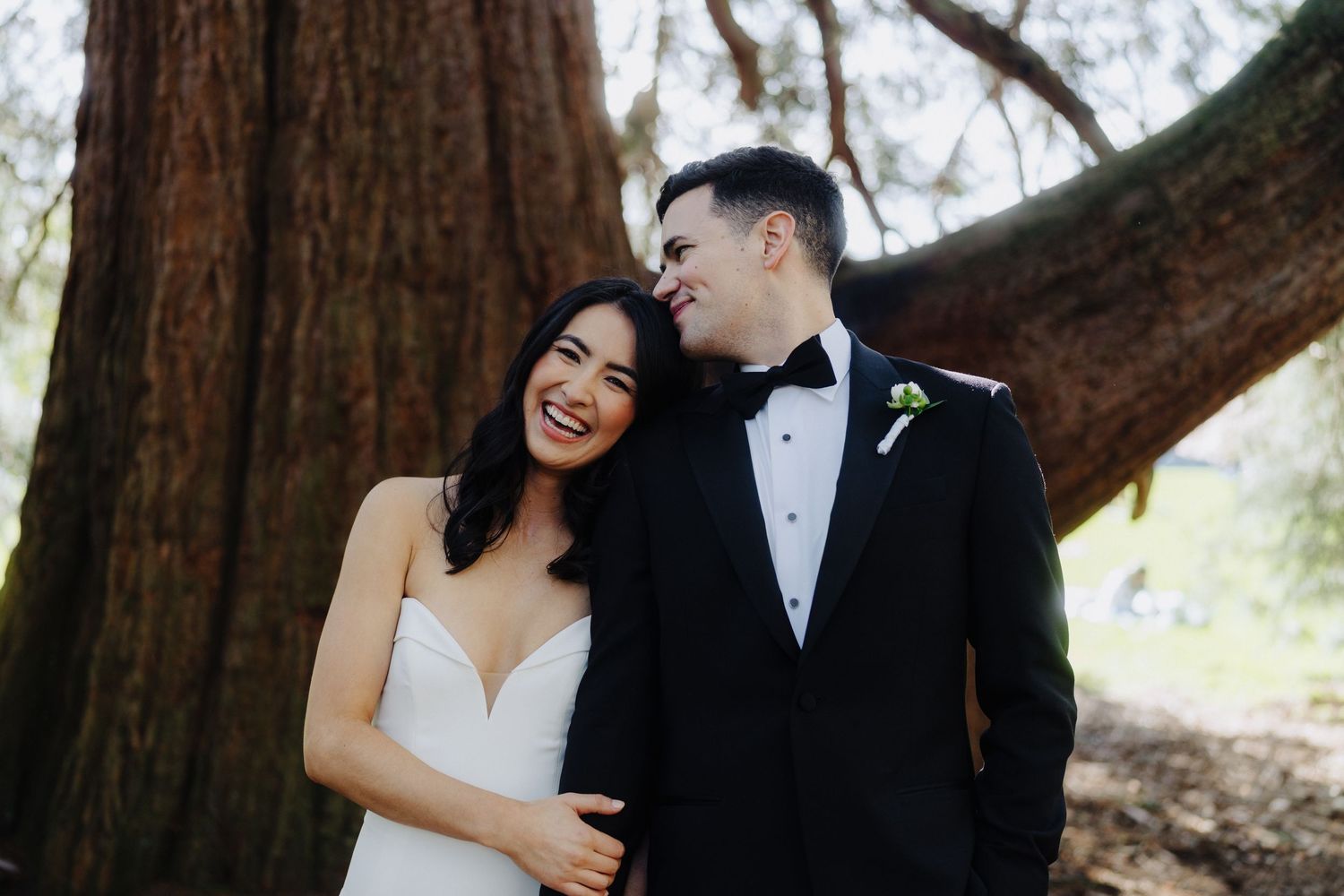 A happy couple sharing a candid moment in formal wedding attire under towering trees.