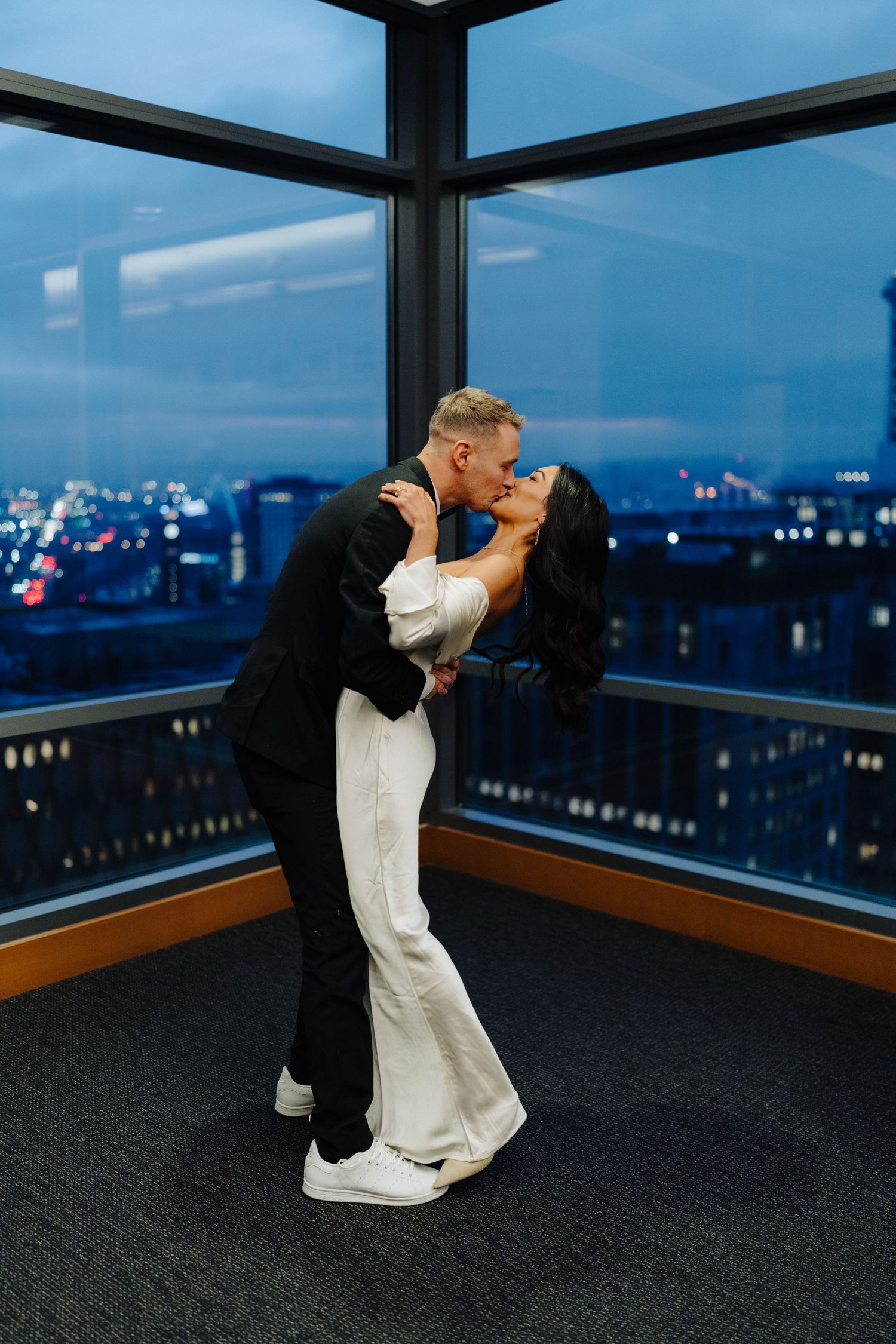 A couple embraces in a romantic dance pose against a panoramic cityscape view through glass windows at night.