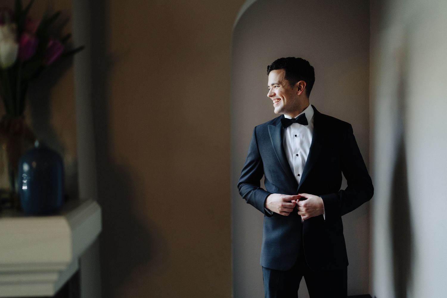 A person in a classic black tuxedo with bow tie stands by a window in soft natural light.