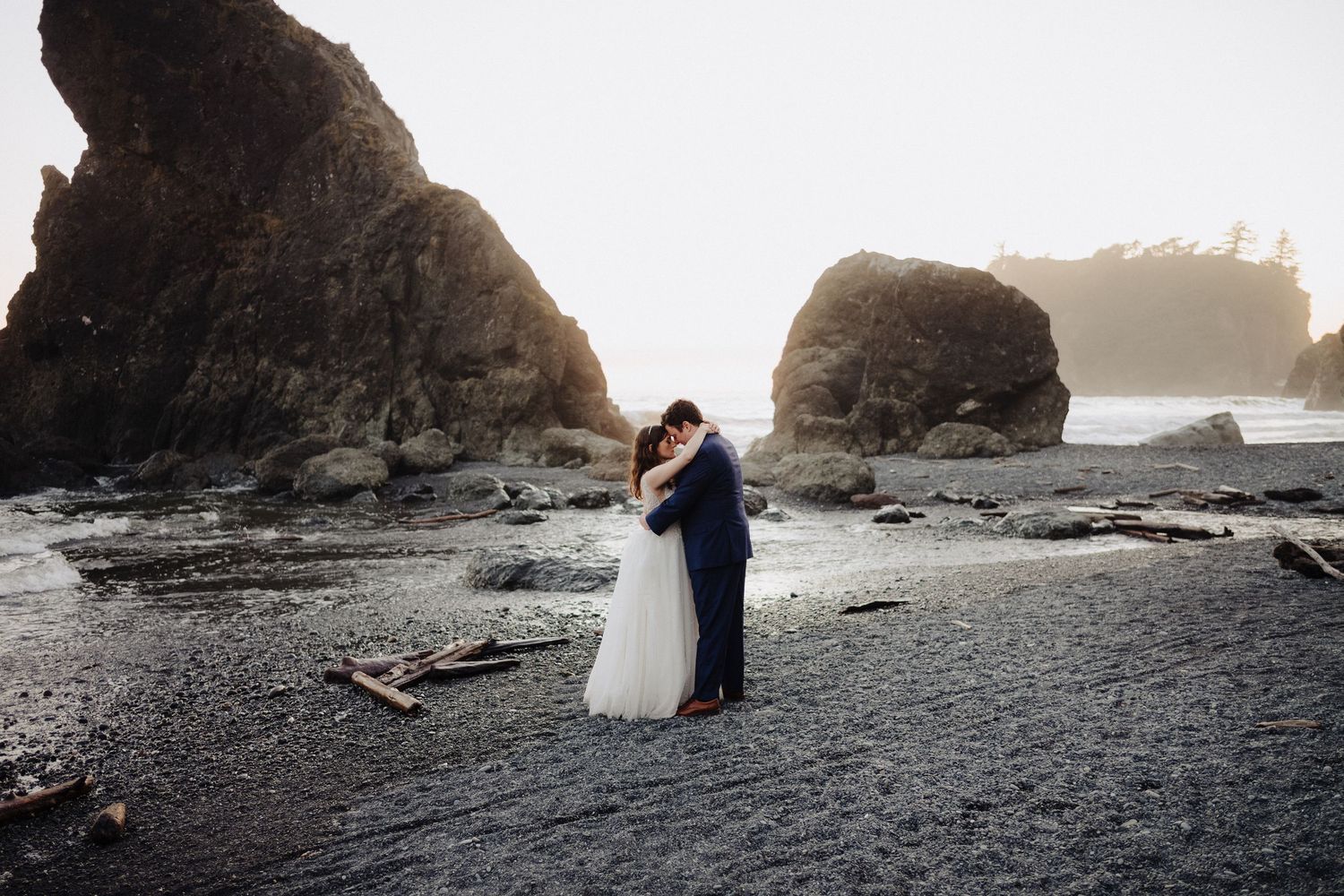Wedding couple standing on dark beach with dramatic rock formations and ocean waves.
