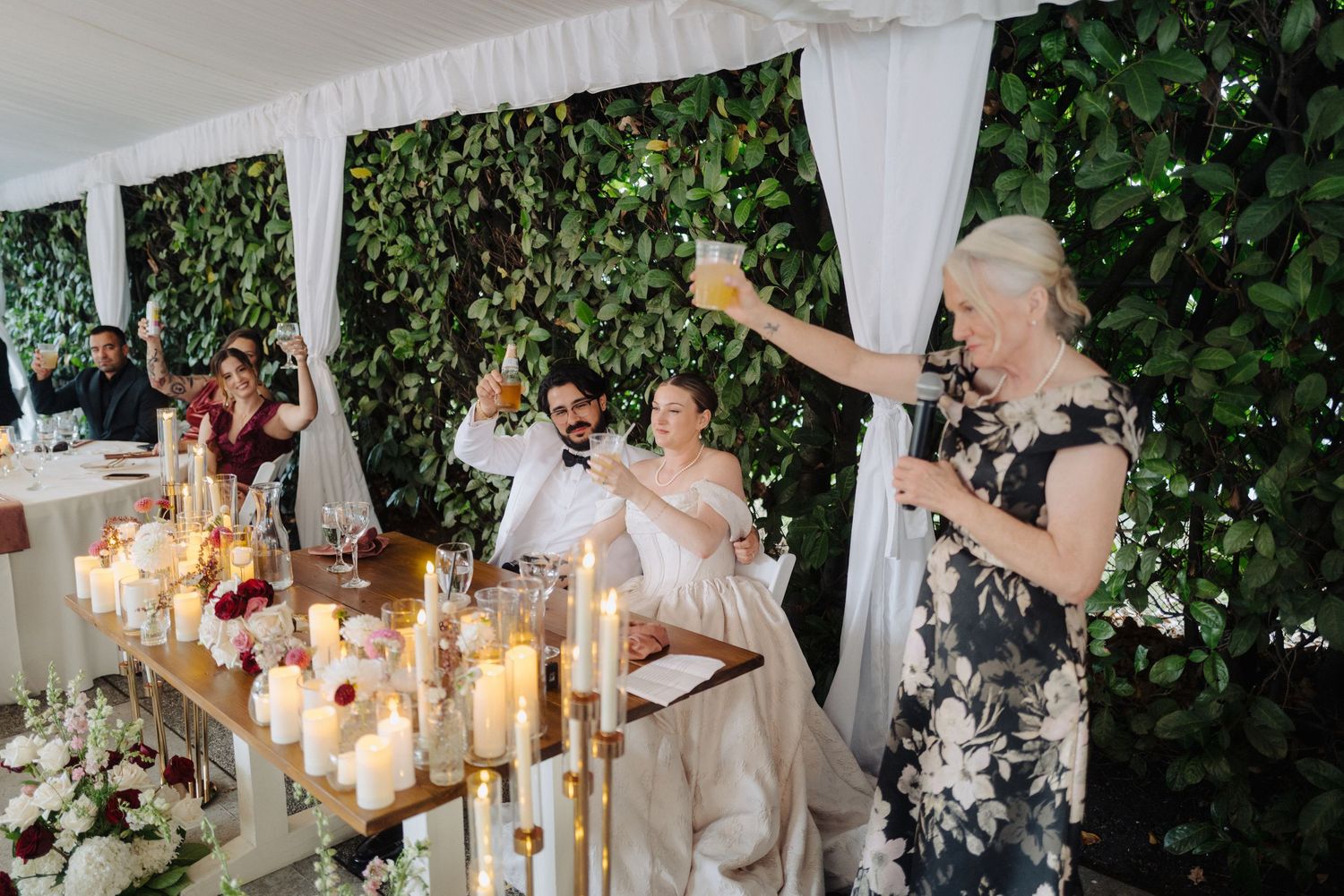 Wedding reception tent decorated with hanging lights and white drapery against green foliage.