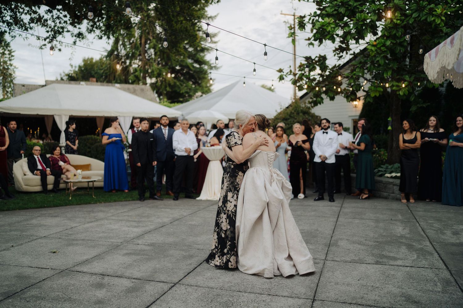Wedding couple dancing surrounded by guests under string lights and white tents at an outdoor reception.