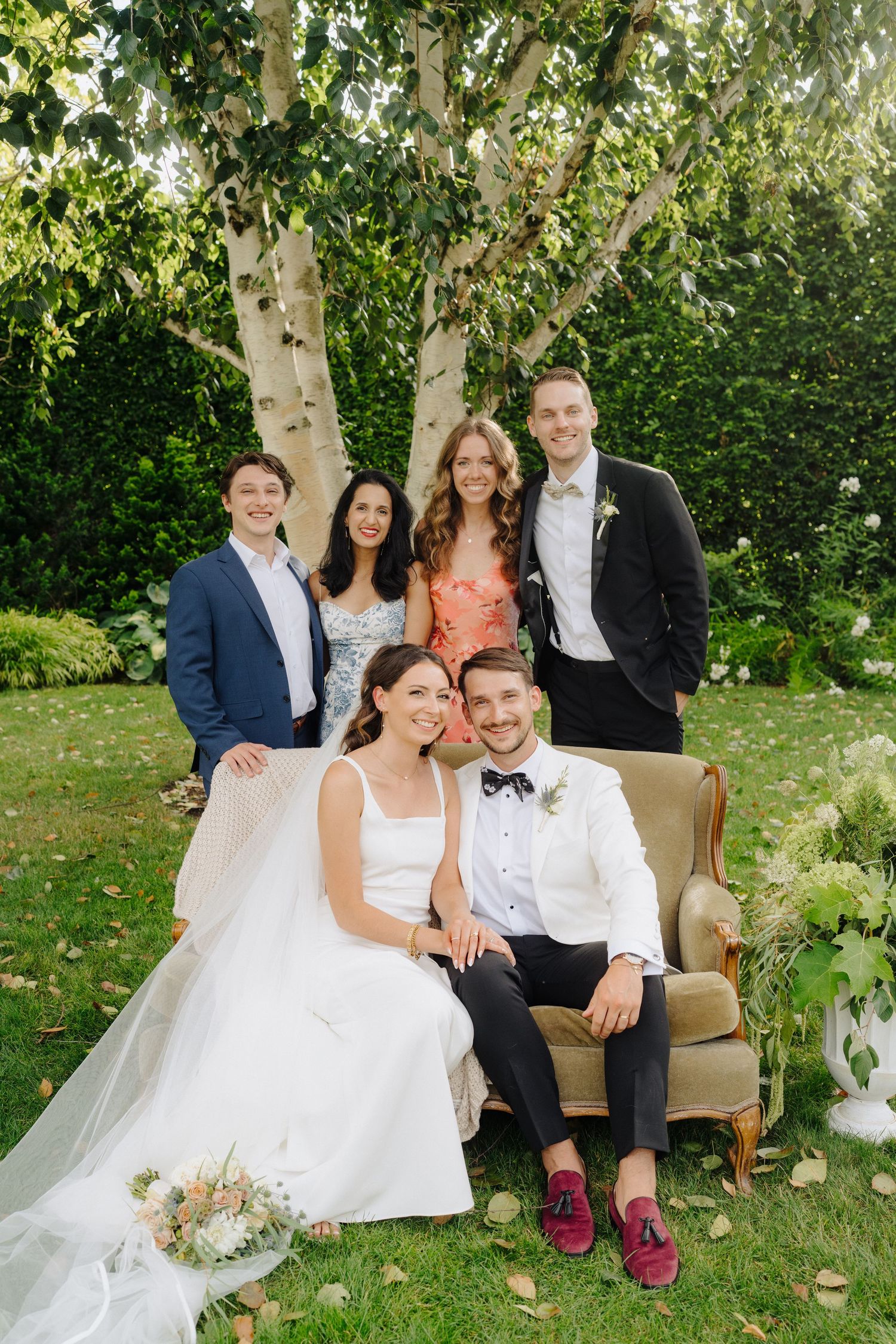 Wedding celebration group poses casually on outdoor furniture under birch trees.