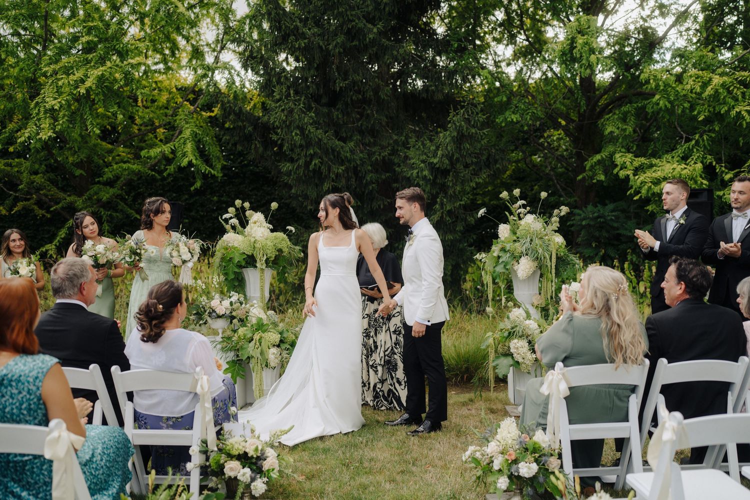 Garden wedding ceremony with guests seated on white chairs surrounded by lush greenery.