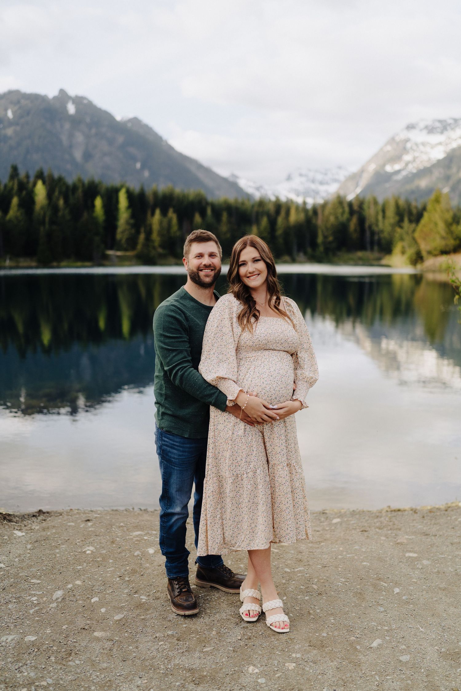 A couple stands together on a rocky lakeshore with snow-capped mountains and evergreen forest in the background.