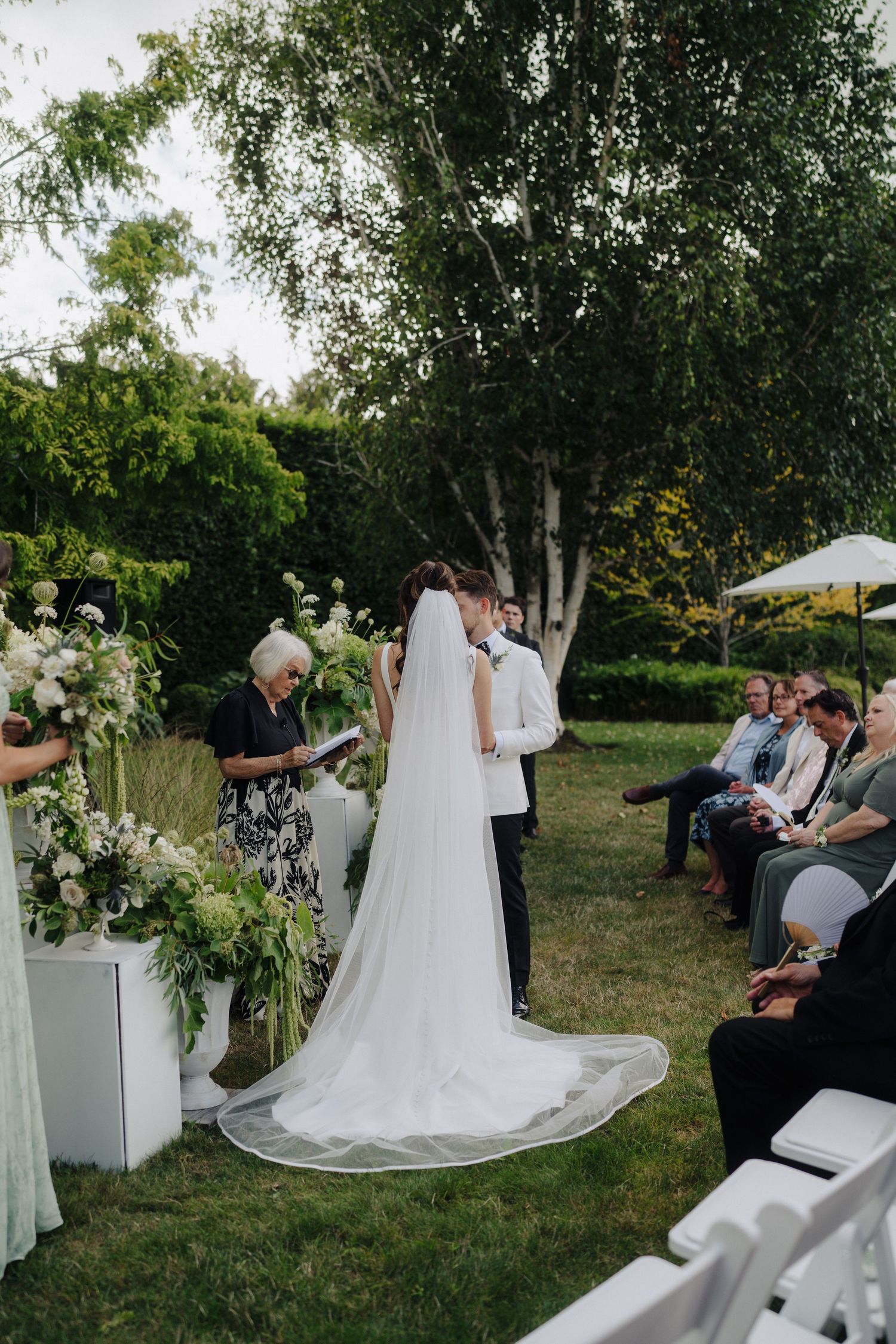 Outdoor wedding ceremony setup with white chairs and floral arrangements under birch trees.