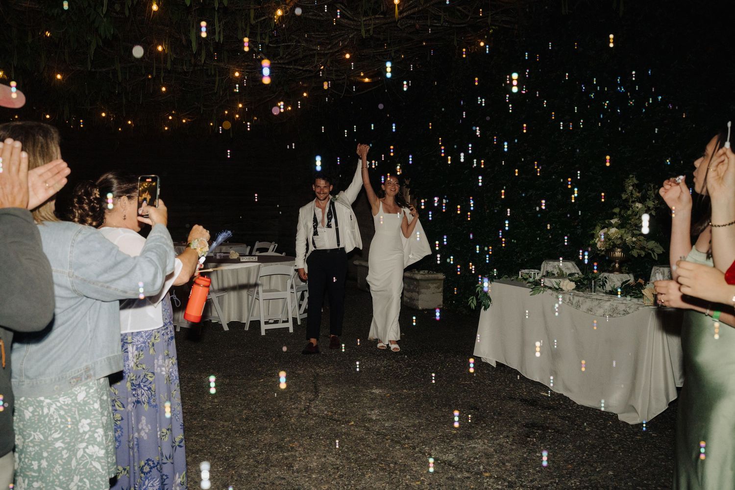 Wedding guests dance under twinkling lights at an evening outdoor reception.