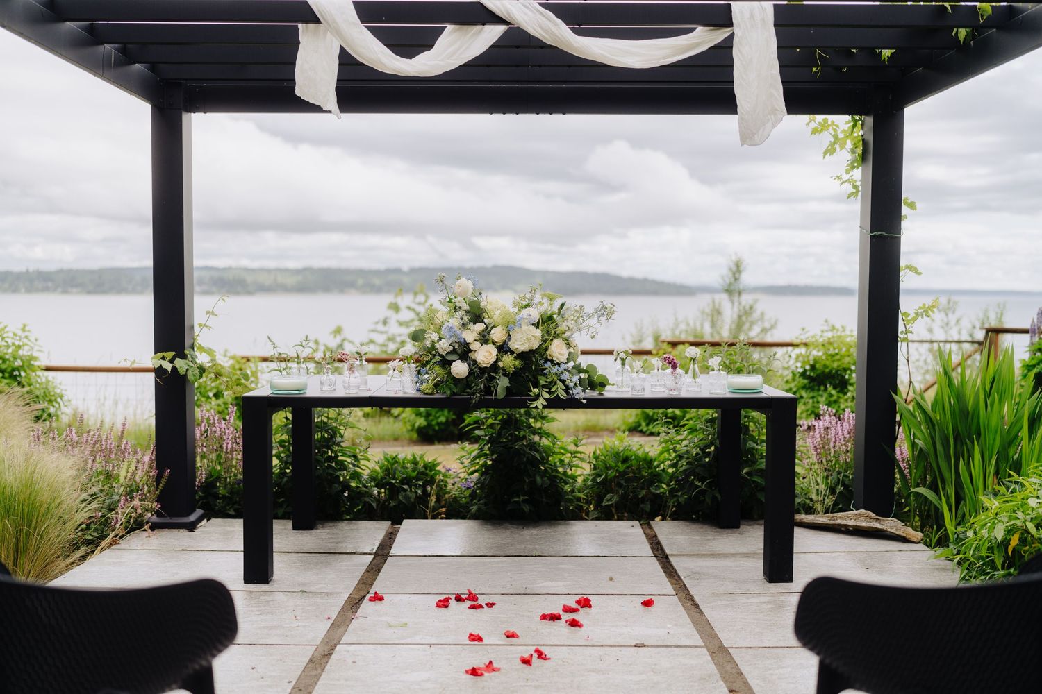 An outdoor wedding ceremony setup with white drapes and rose petals leading to a waterfront view.