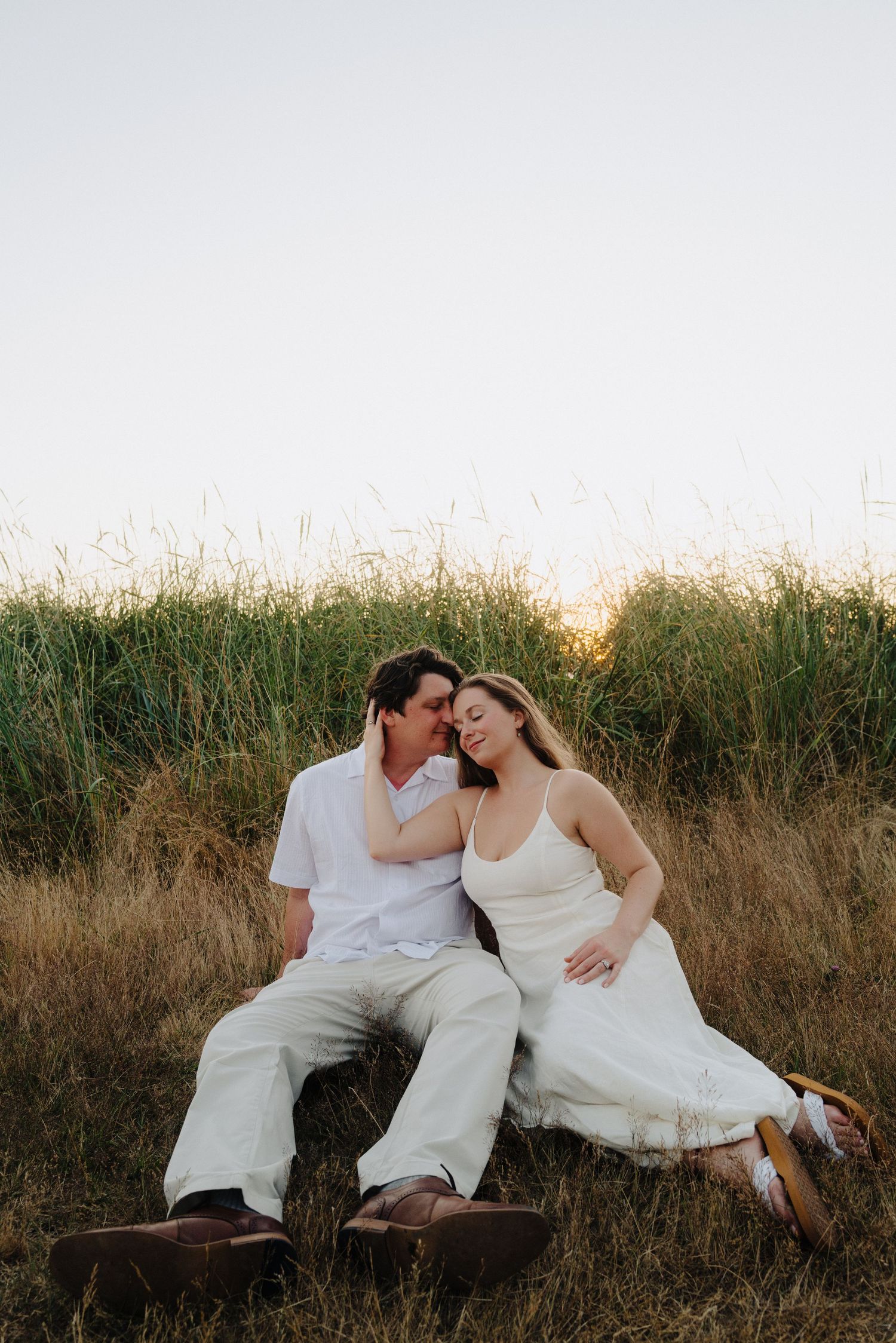 A couple in white clothing reclines together among tall grass during golden hour.