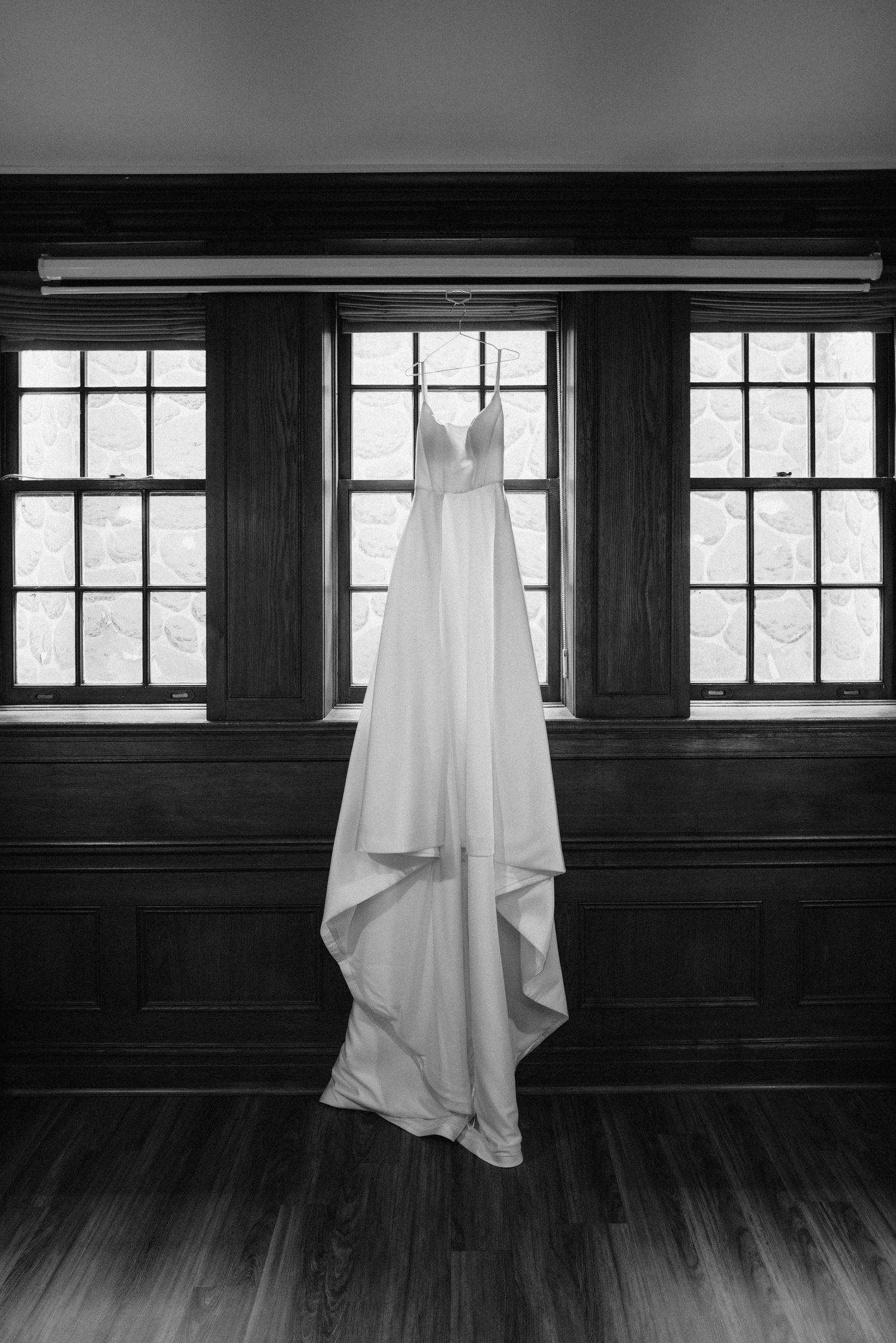 A white wedding dress hangs elegantly against dark wood paneling and three large windows.