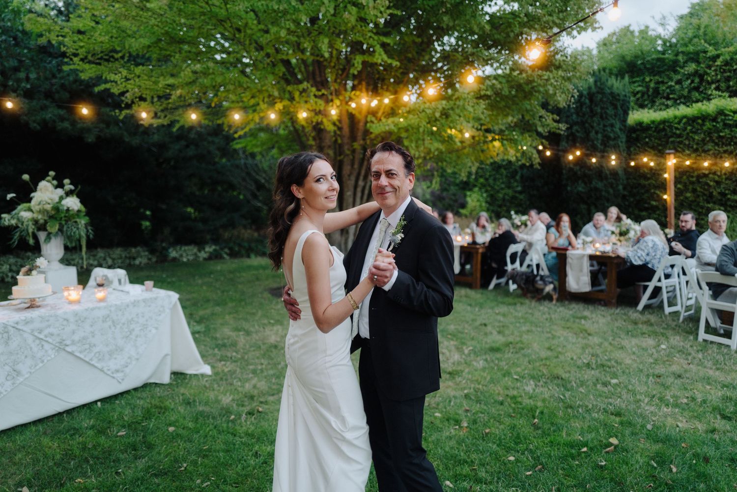 String lights illuminate an outdoor evening wedding reception on a green lawn.