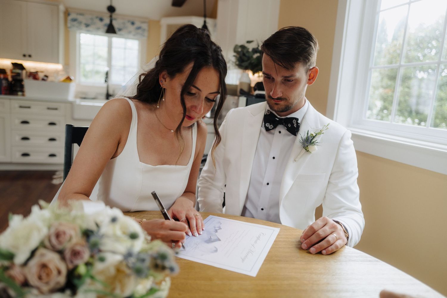 A couple signing marriage documents at a wooden table with a floral bouquet nearby.