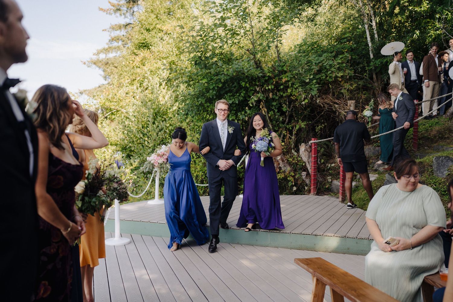 Outdoor wedding ceremony processional on wooden deck with attendants in blue and purple dresses.