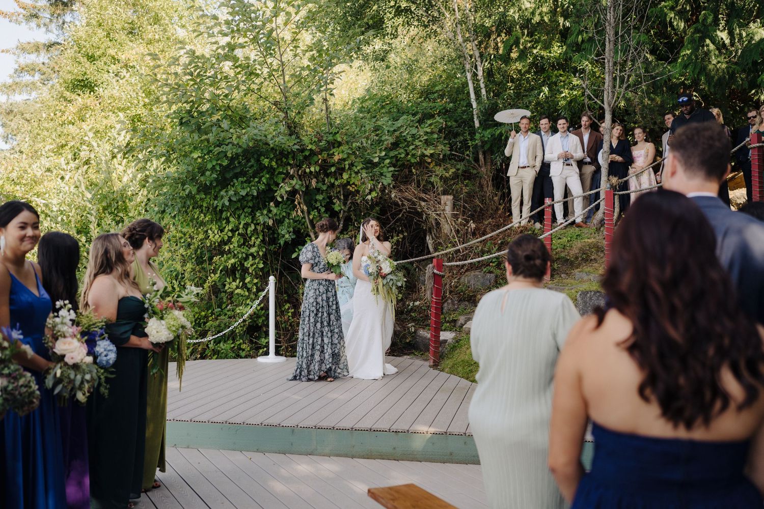 Wedding guests gather outdoors under trees during a daytime celebration.
