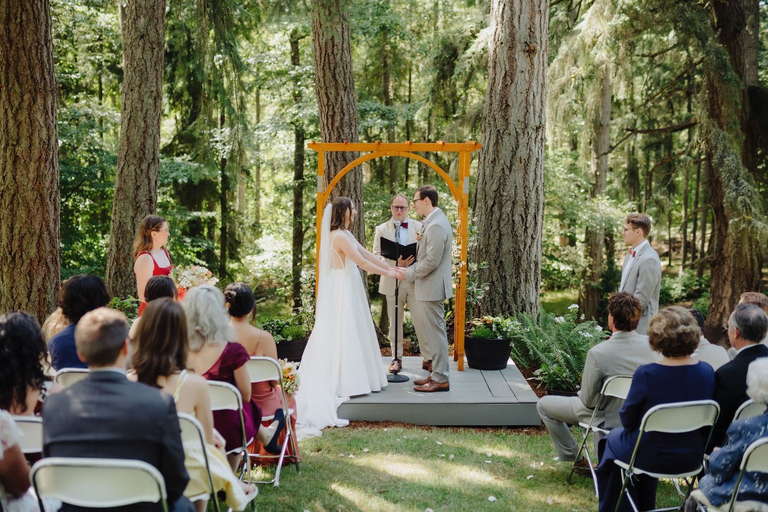An intimate forest wedding ceremony with guests seated on folding chairs among tall trees.