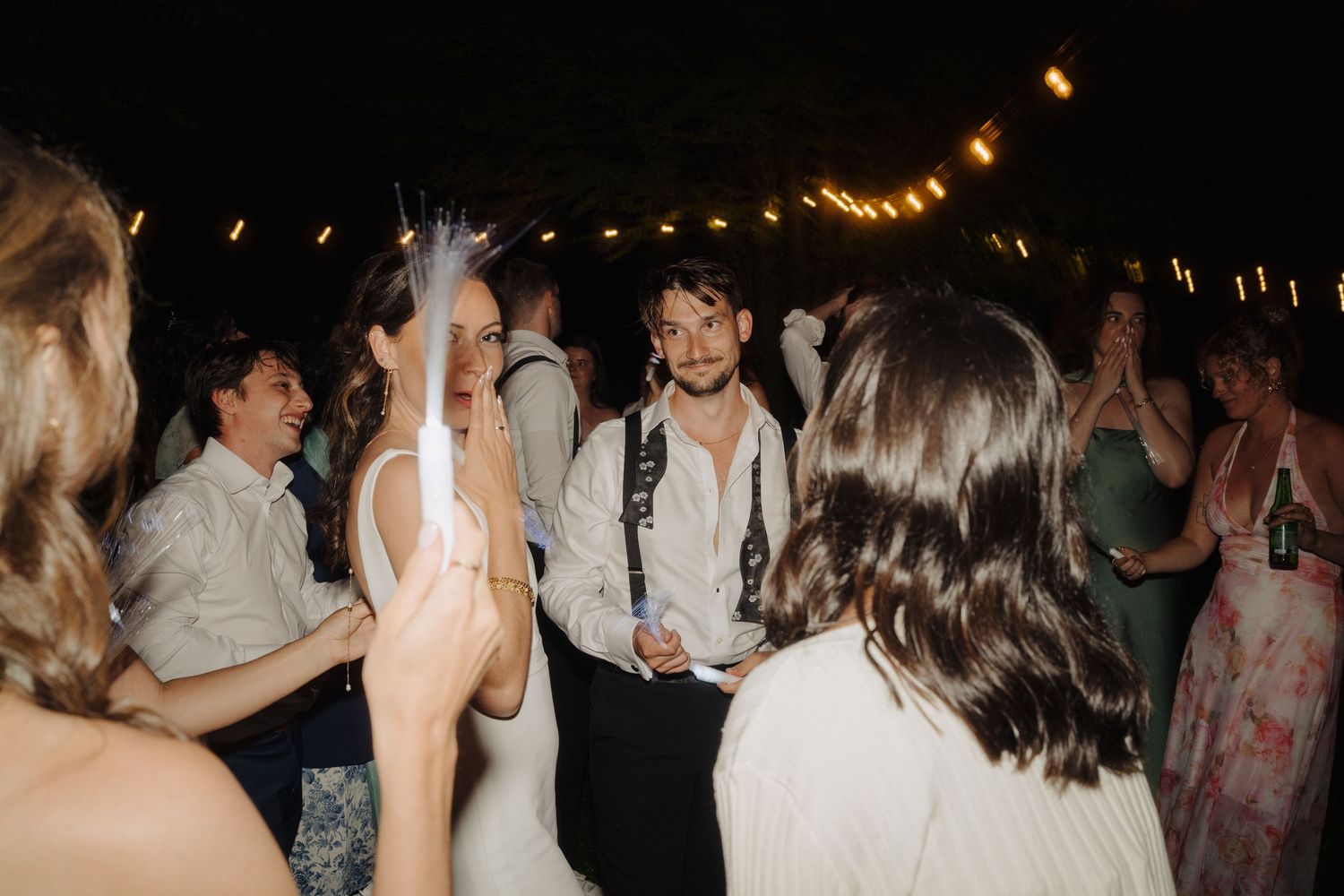 Wedding reception guests dancing and celebrating on a dark dance floor under string lights.