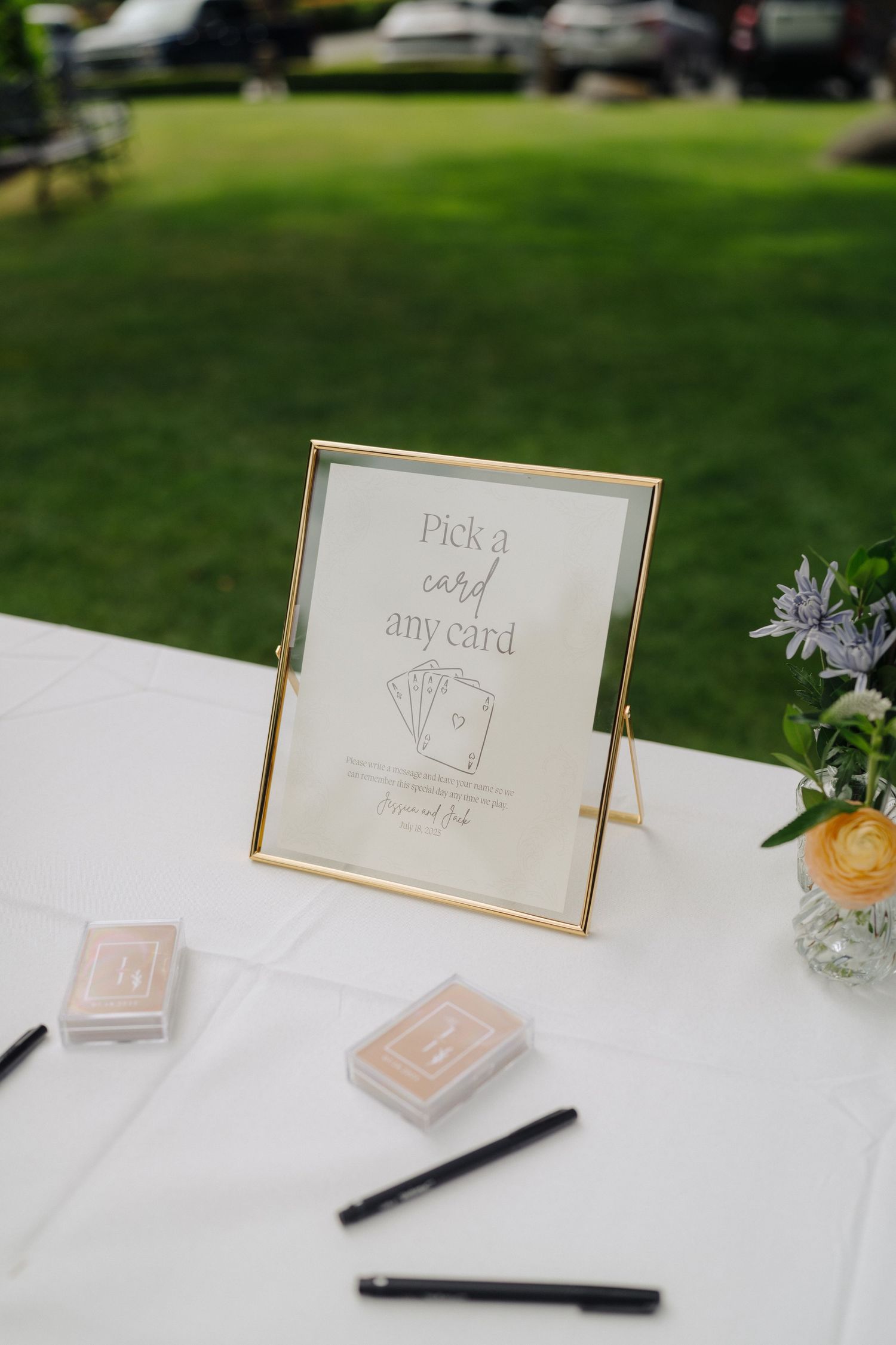 Gold-framed 'Pick a card' sign with playing cards on white tablecloth at wedding reception.