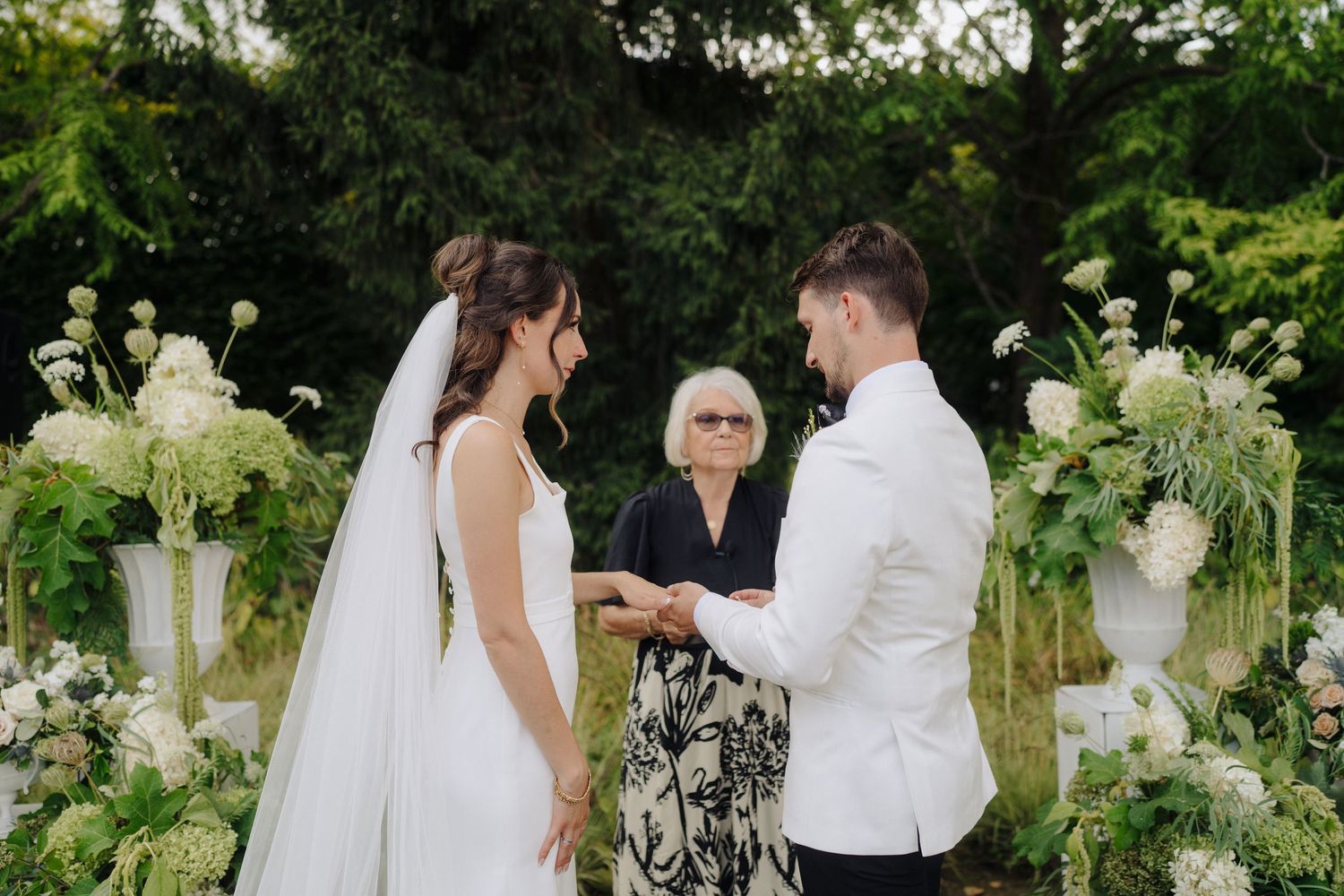 Couple exchanges rings during outdoor garden wedding ceremony surrounded by white floral arrangements.