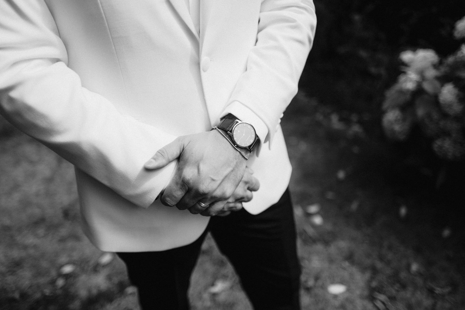 Close-up detail of groom's white suit jacket and watch against garden background.