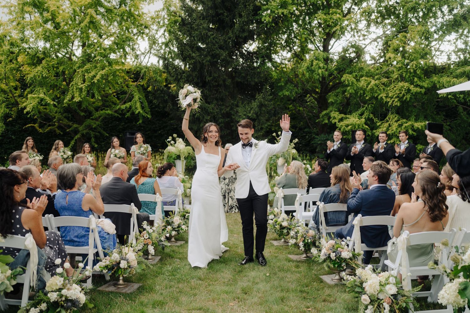 Wedding celebration in a garden setting with rows of white chairs and natural greenery.