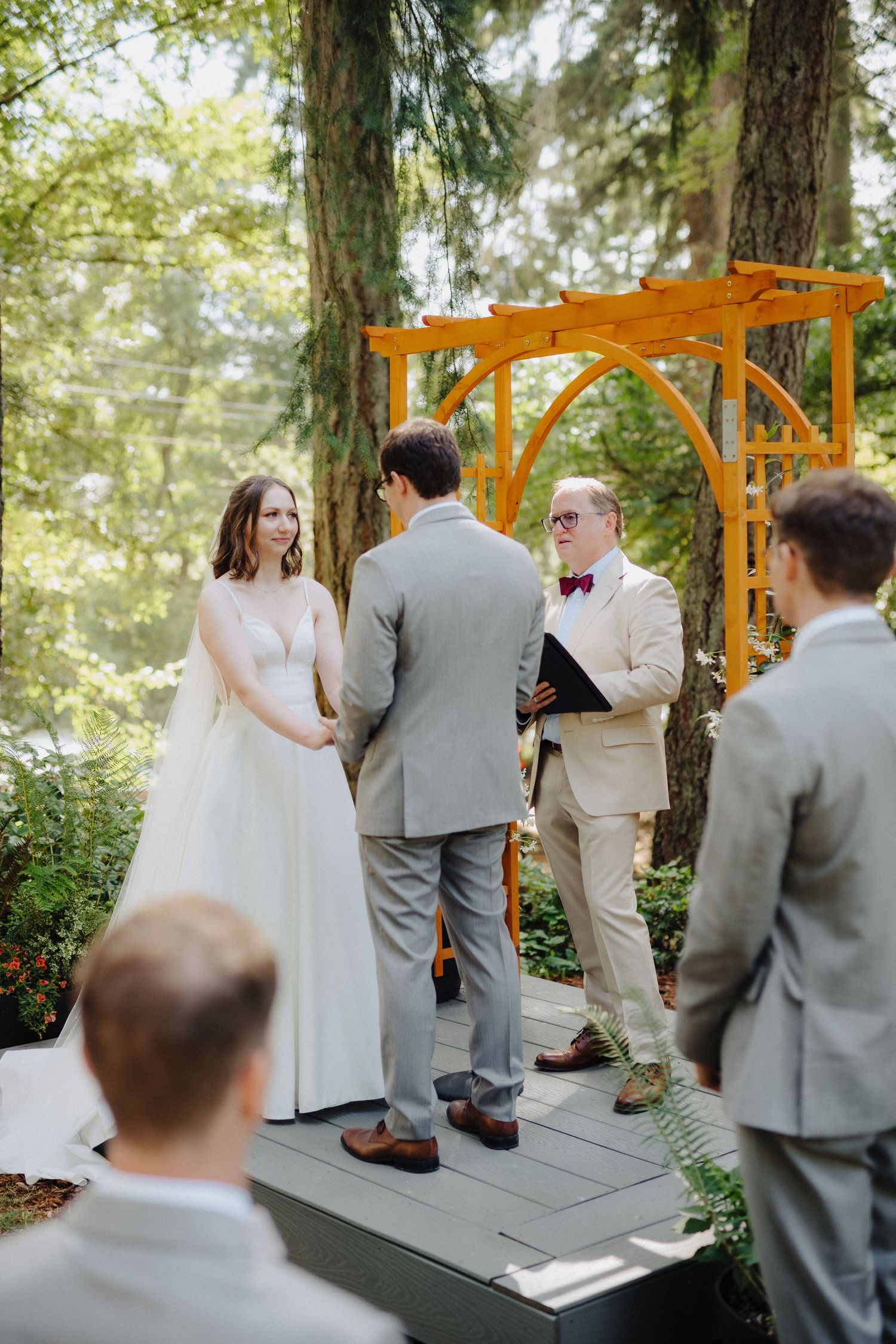 Outdoor wedding ceremony under tall trees with wooden arch and guests in grey suits.