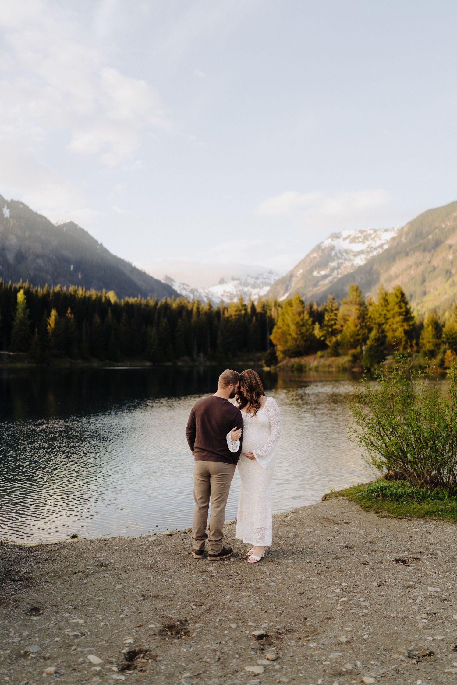 Two people stand together on a gravel shoreline overlooking a pristine mountain lake at sunset.