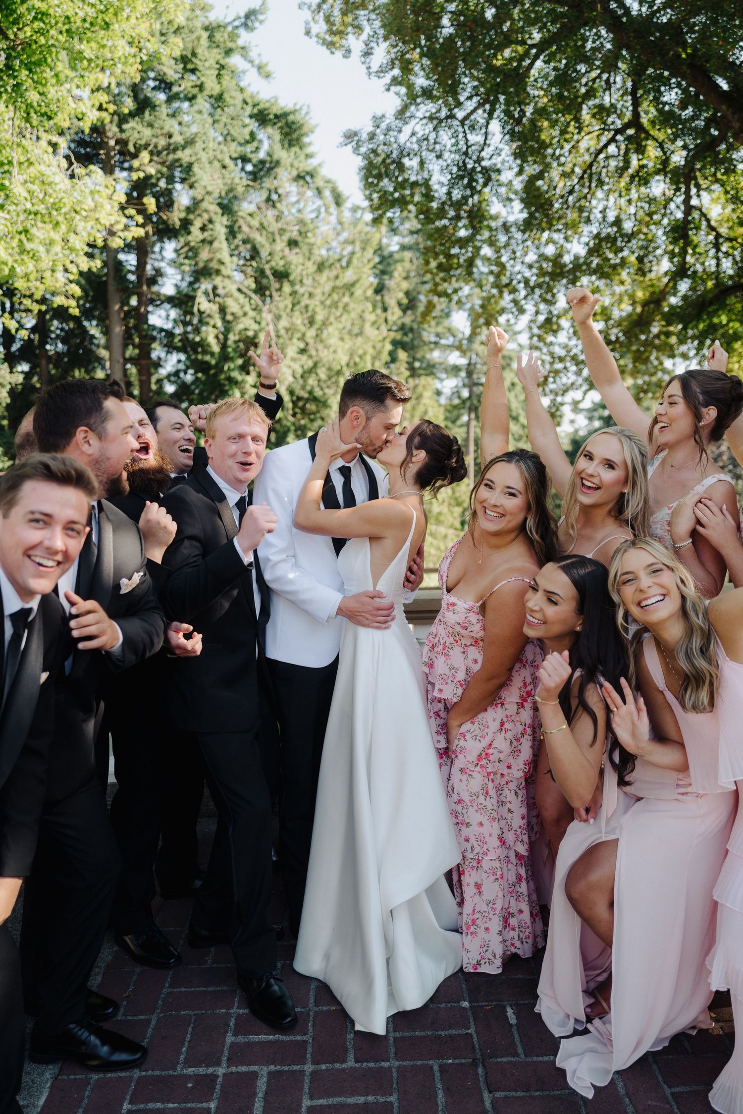 Wedding party celebrates joyfully outdoors with the newlyweds under trees.