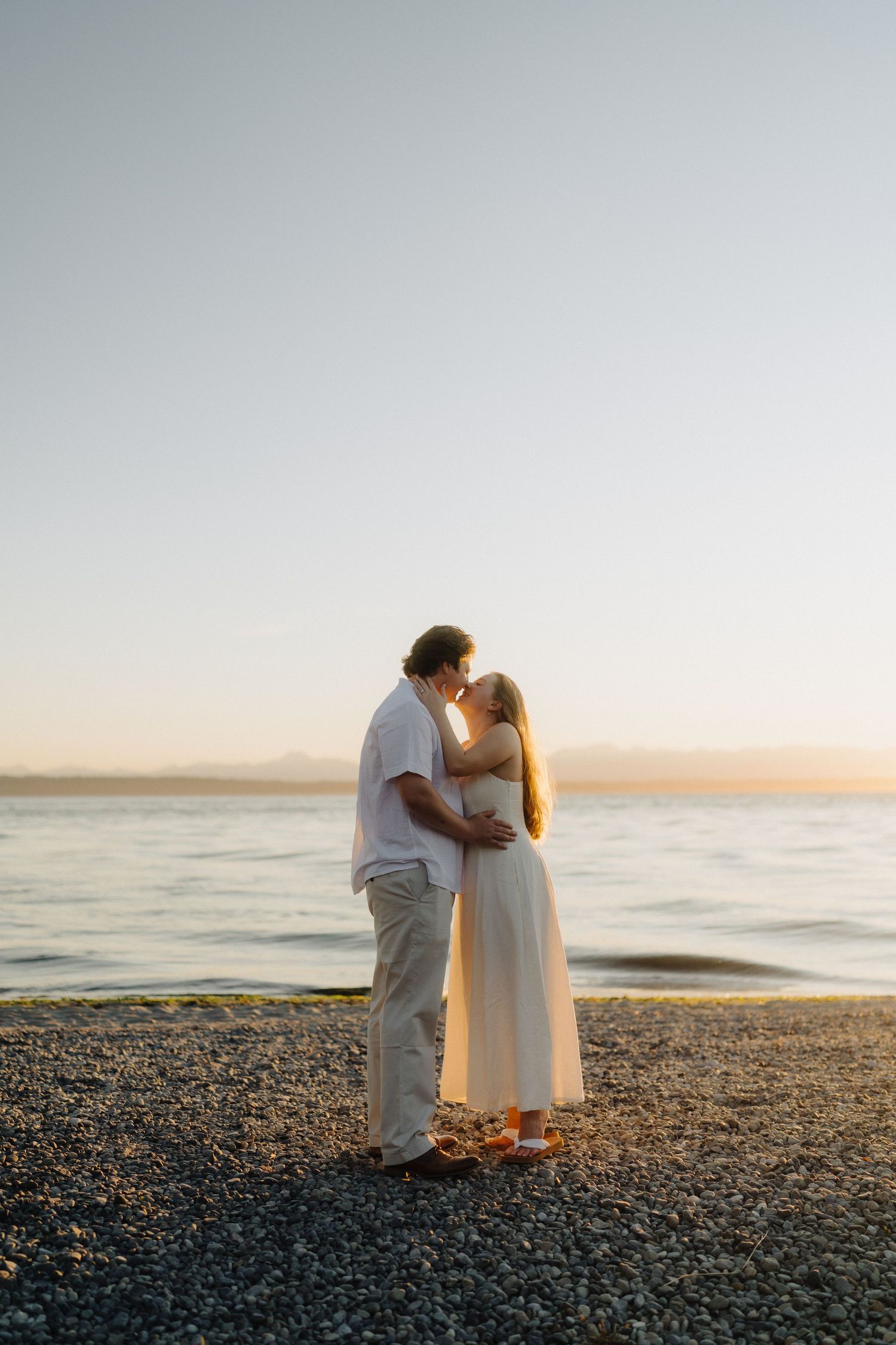 A couple shares a romantic moment on a rocky beach at sunset with the ocean in the background.