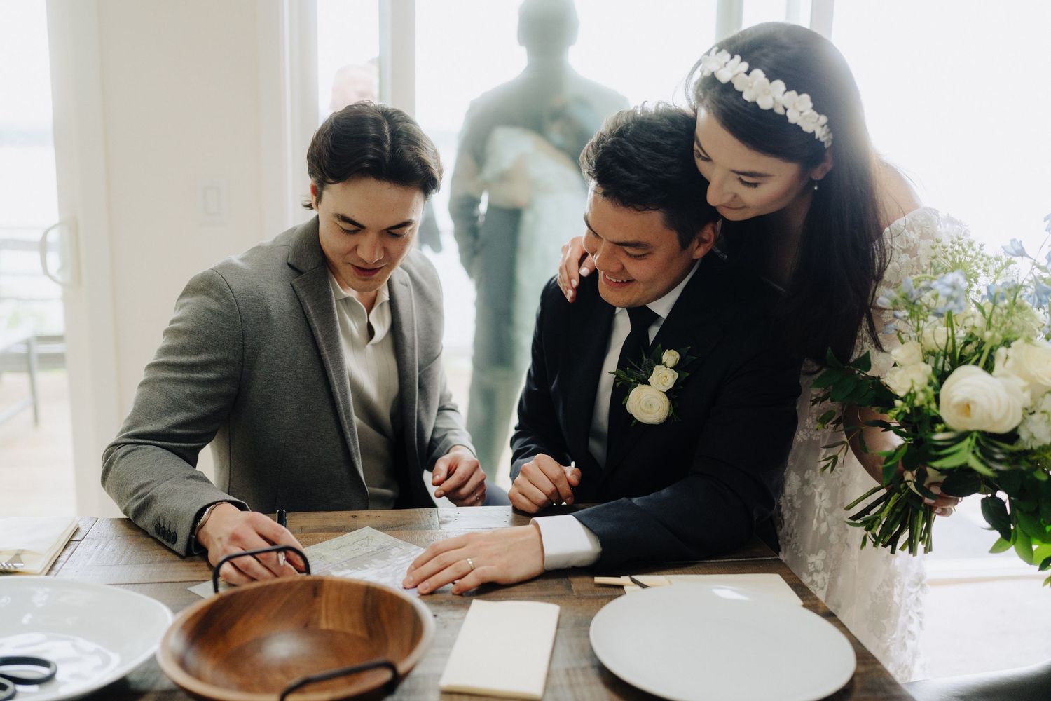 Wedding couple signing marriage documents at wooden table with floral arrangements nearby.