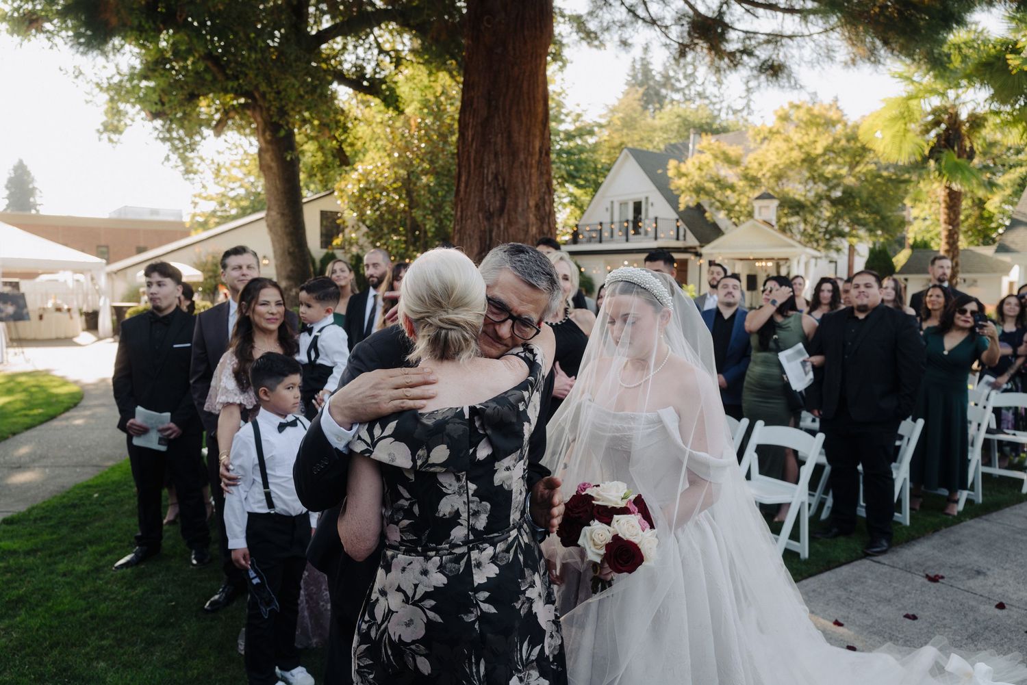 Wedding ceremony in progress with guests seated on white chairs under tall trees.