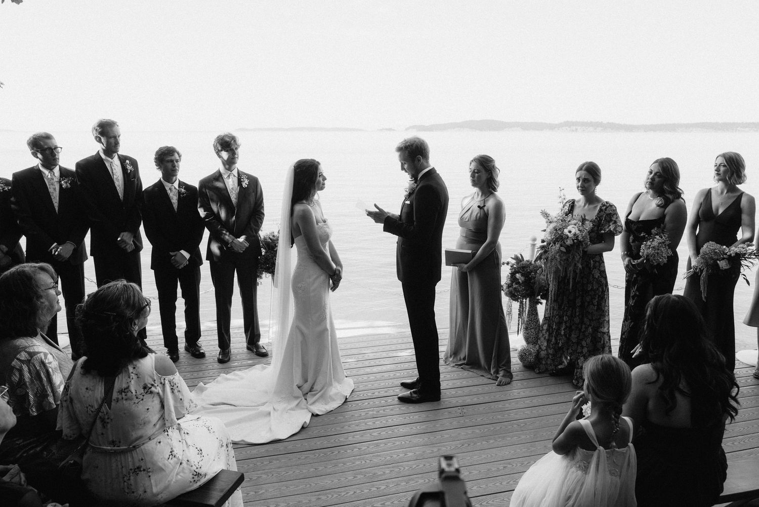 Black and white photo of a wedding ceremony on a waterfront deck with guests watching.