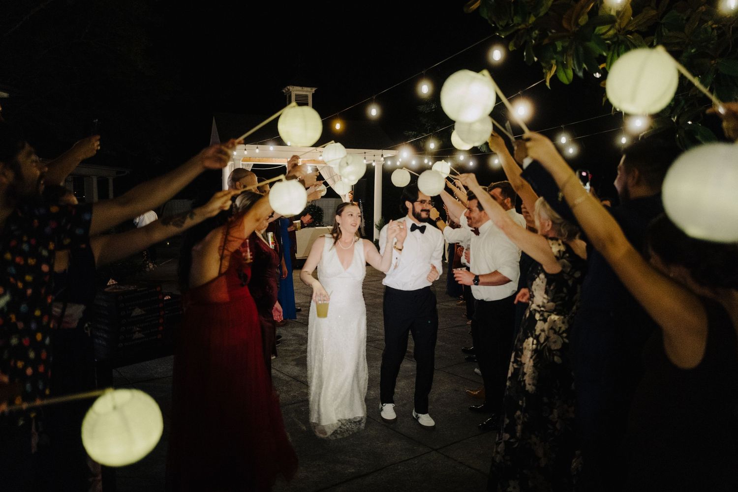Paper lanterns illuminate an outdoor wedding reception as guests dance under the evening sky.