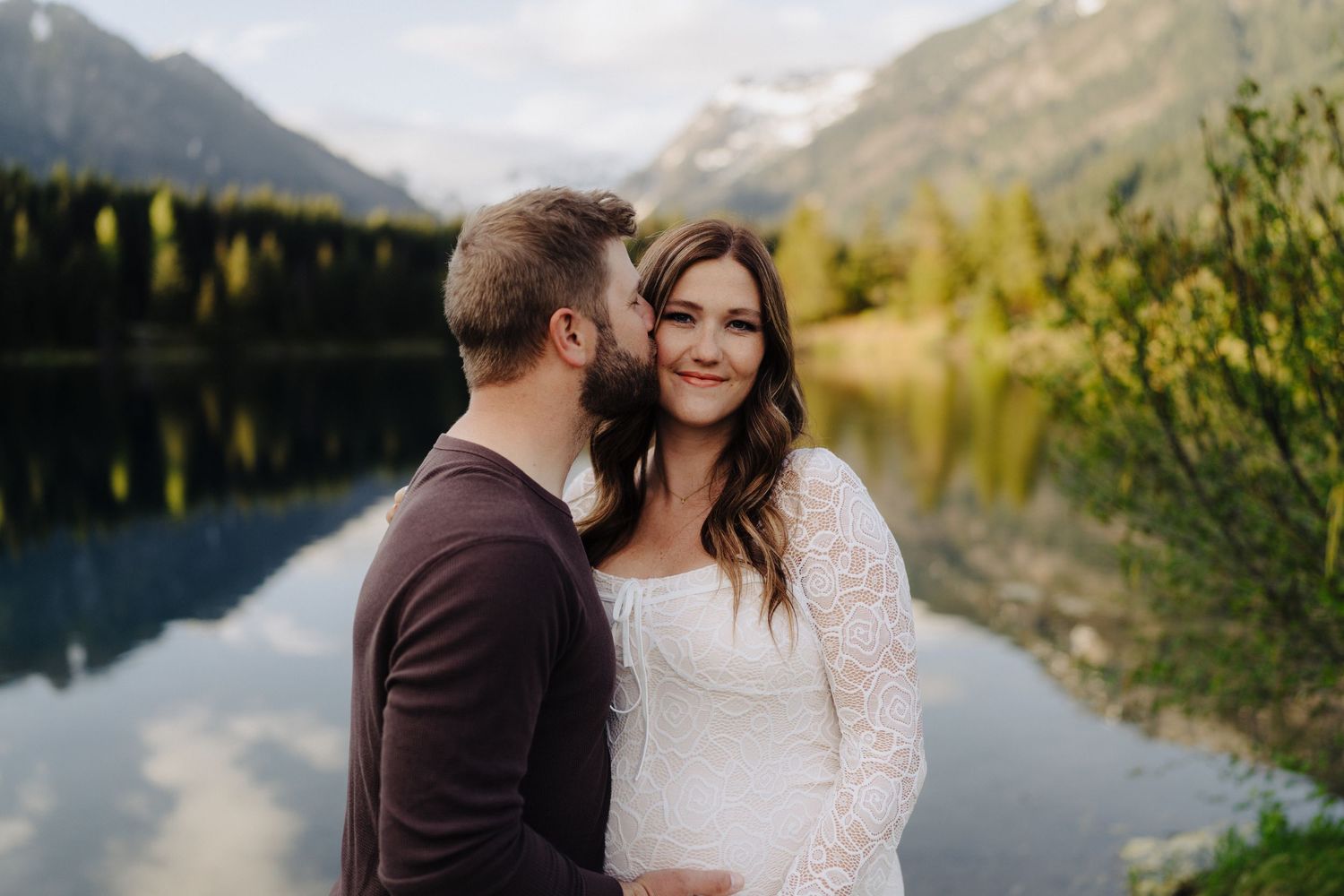 Couple shares tender moment with scenic mountain lake and forest landscape behind them.