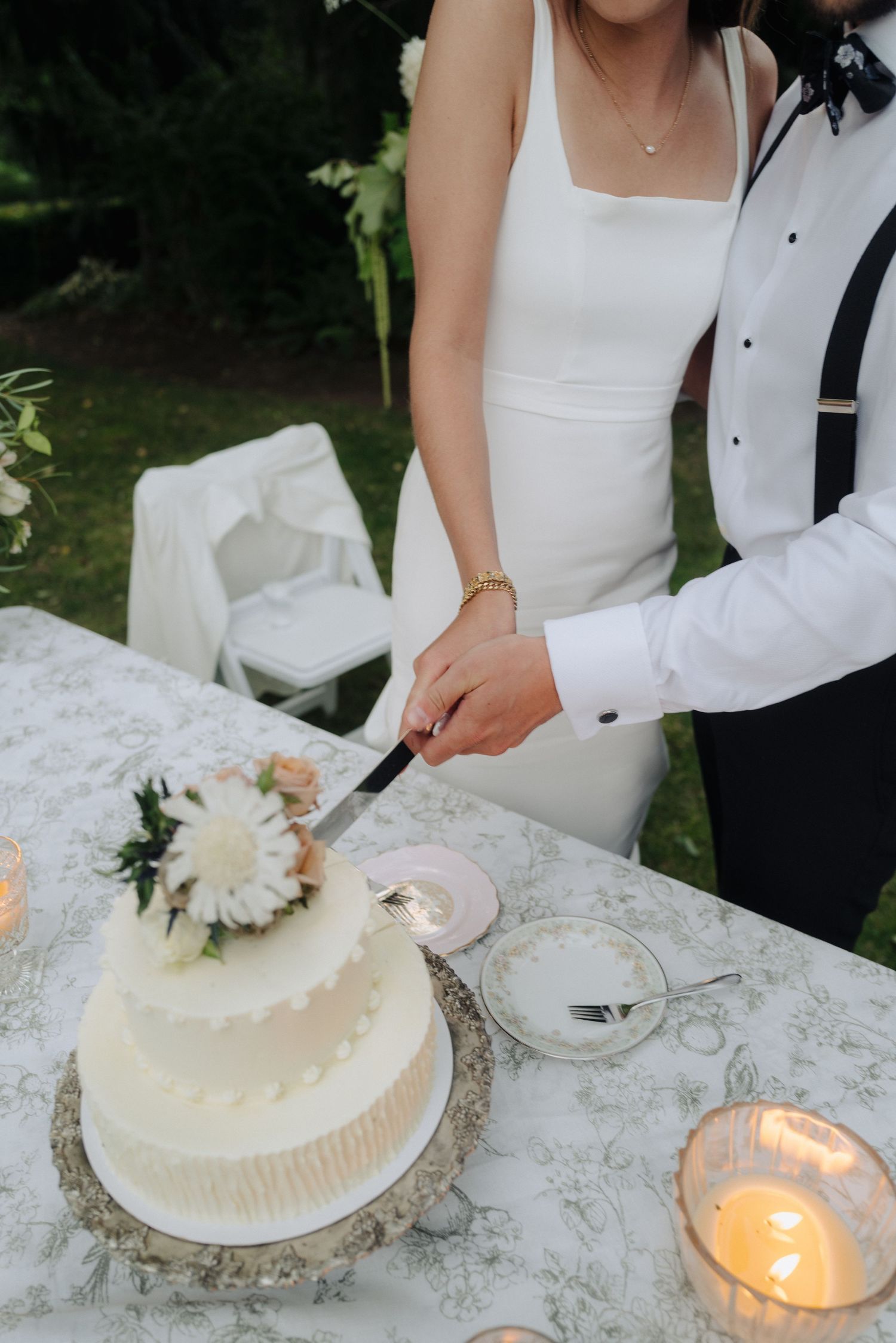 Newlyweds cut into their elegant white wedding cake adorned with flowers on a lace-covered table with glowing candles.