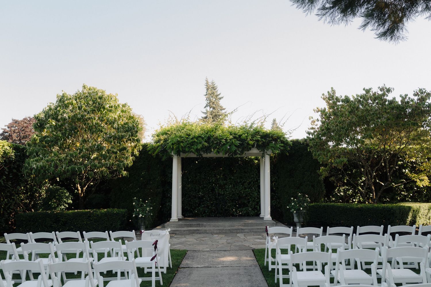 White chairs arranged for an outdoor wedding ceremony under a white pergola surrounded by lush greenery.