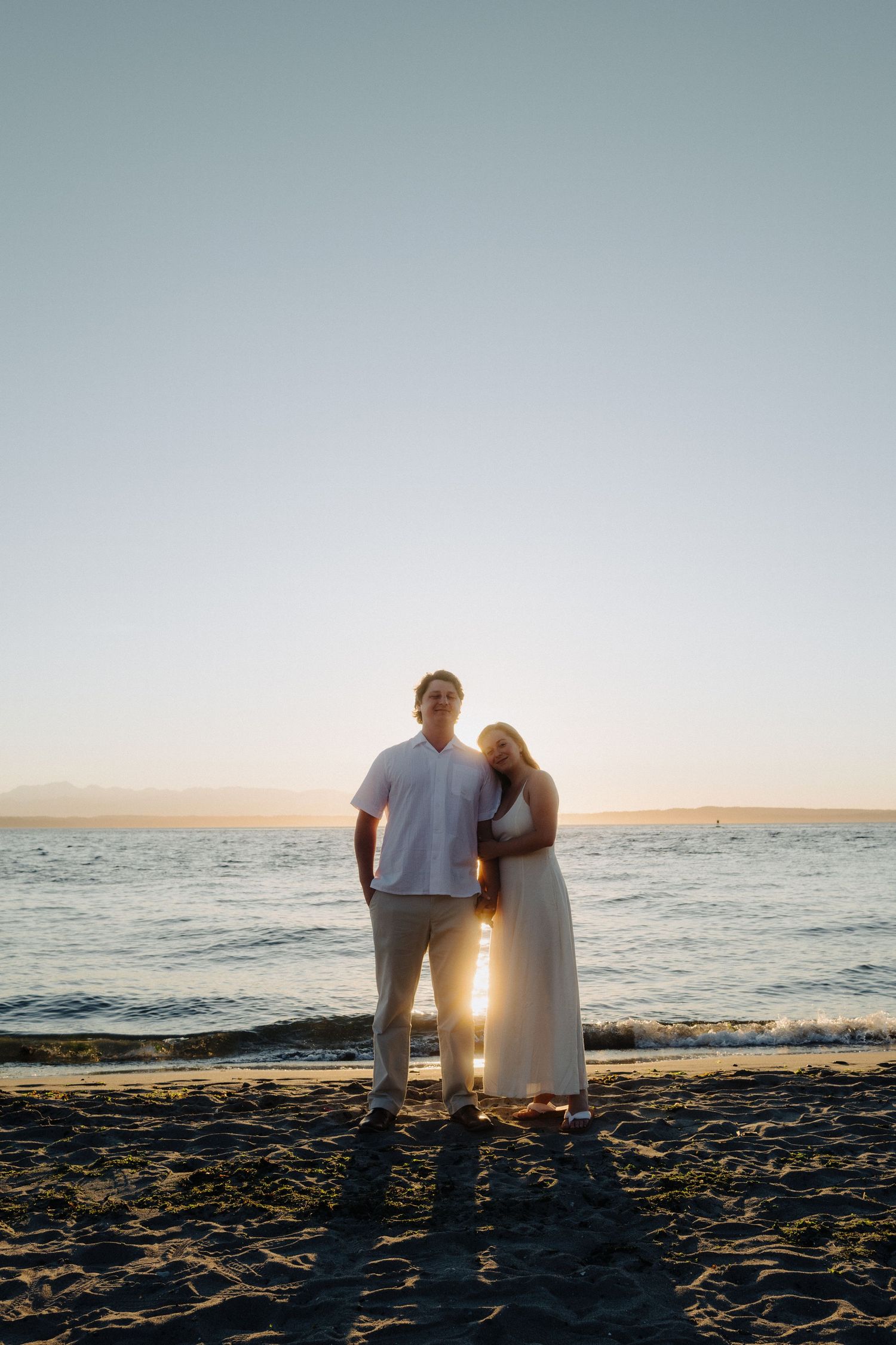 A backlit couple standing together on the beach at sunset with the ocean behind them.