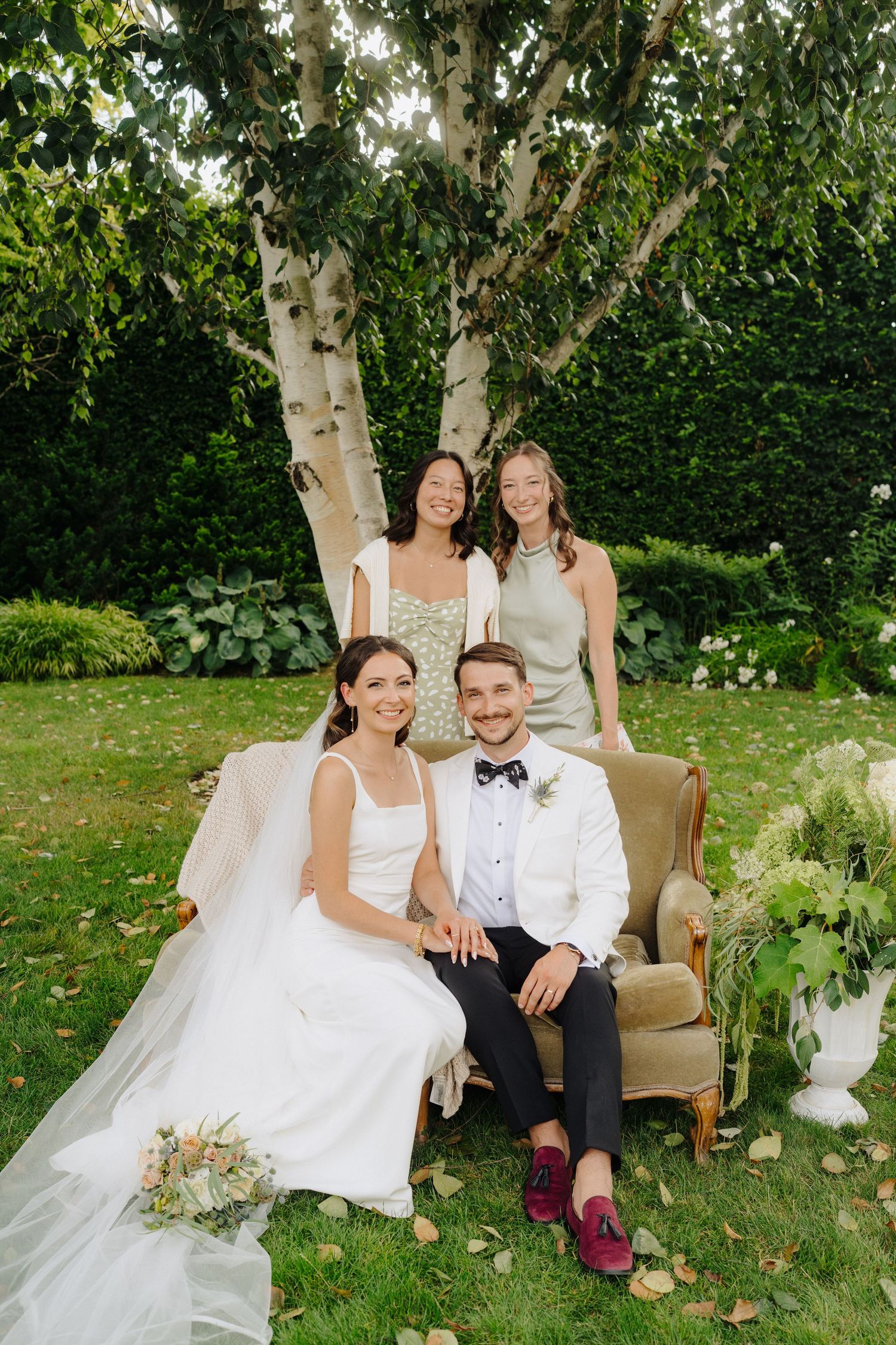 Wedding party relaxes on vintage furniture in a lush garden setting.