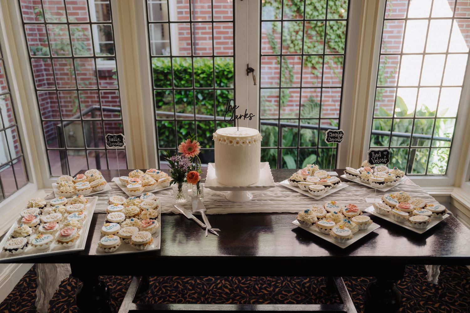 An elegant dessert table display with a white wedding cake and various treats by a bay window.