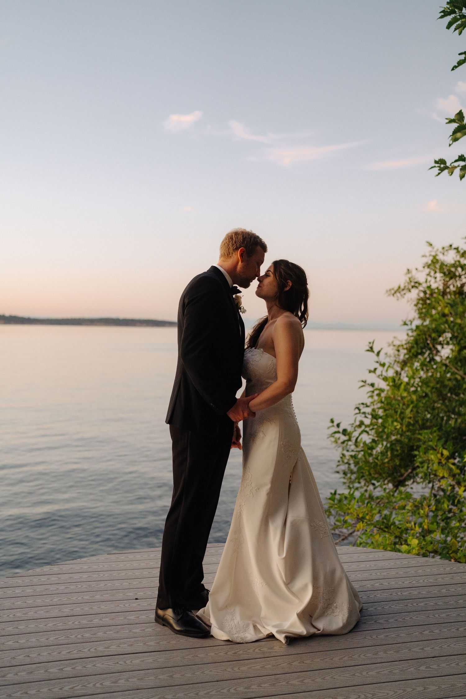 Romantic wedding couple sharing a moment on wooden deck overlooking serene waterfront at sunset.