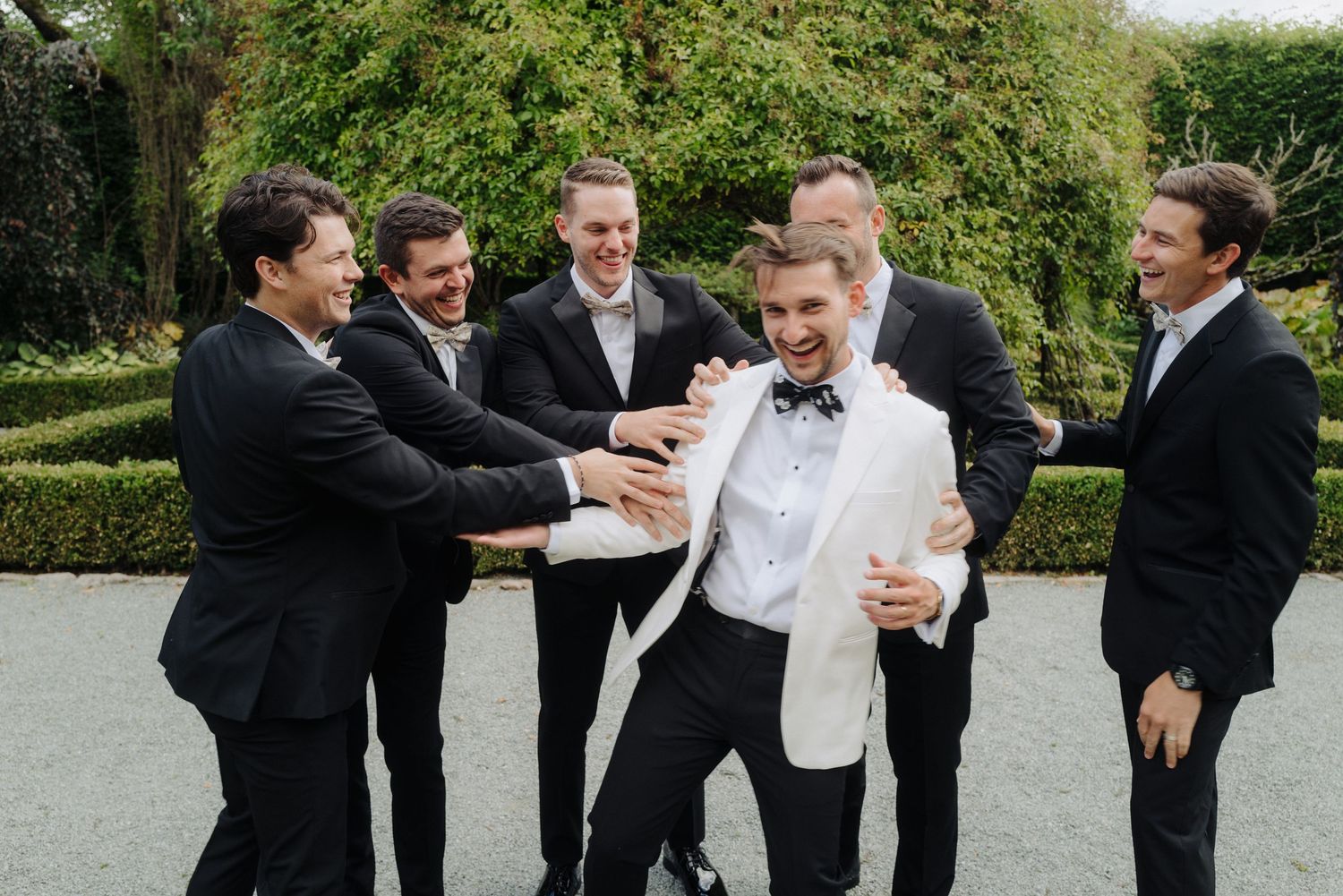 Groomsmen in black suits share candid moments and laughter in a garden.