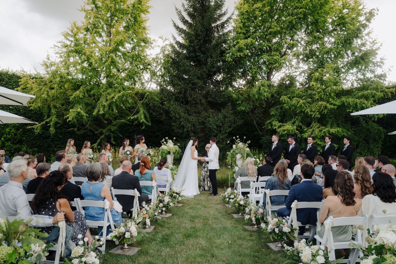 Outdoor wedding ceremony with guests seated on white chairs surrounded by lush greenery.