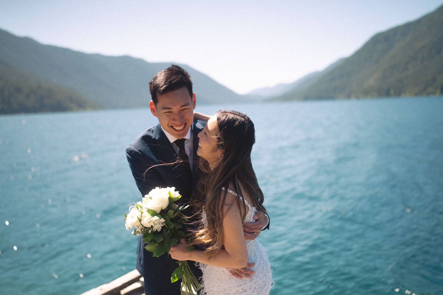 Smiling newlyweds with white bouquet embrace on a boat with turquoise mountain lake backdrop.