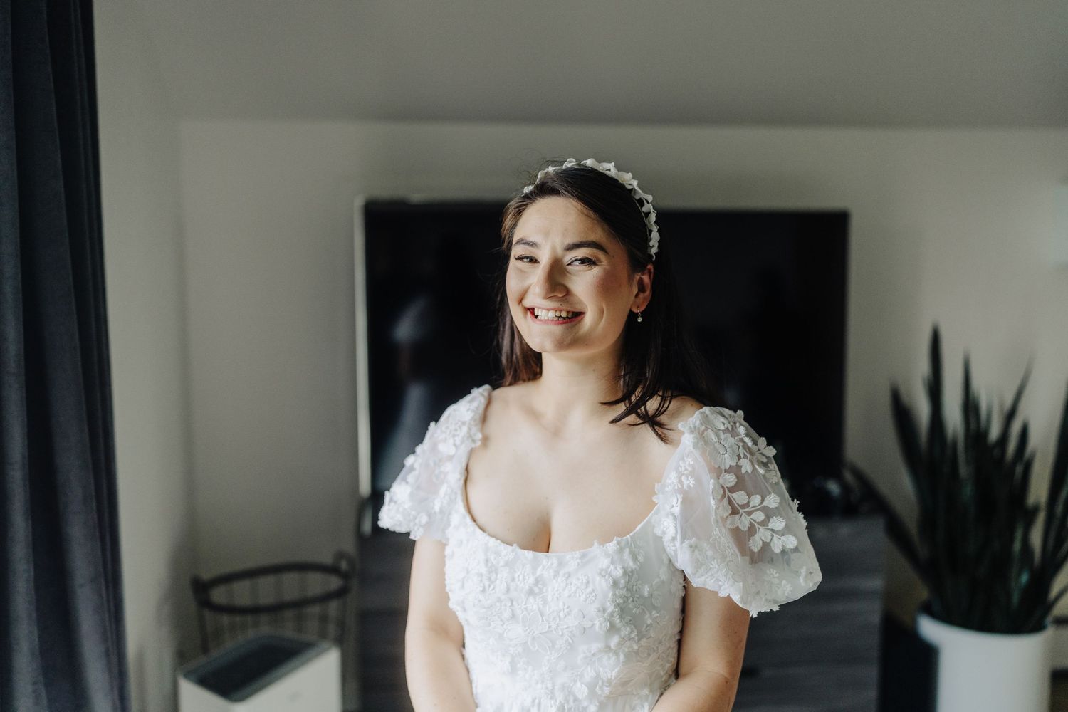 A bride wearing a white dress with puffy sleeves smiles while standing indoors near a houseplant.