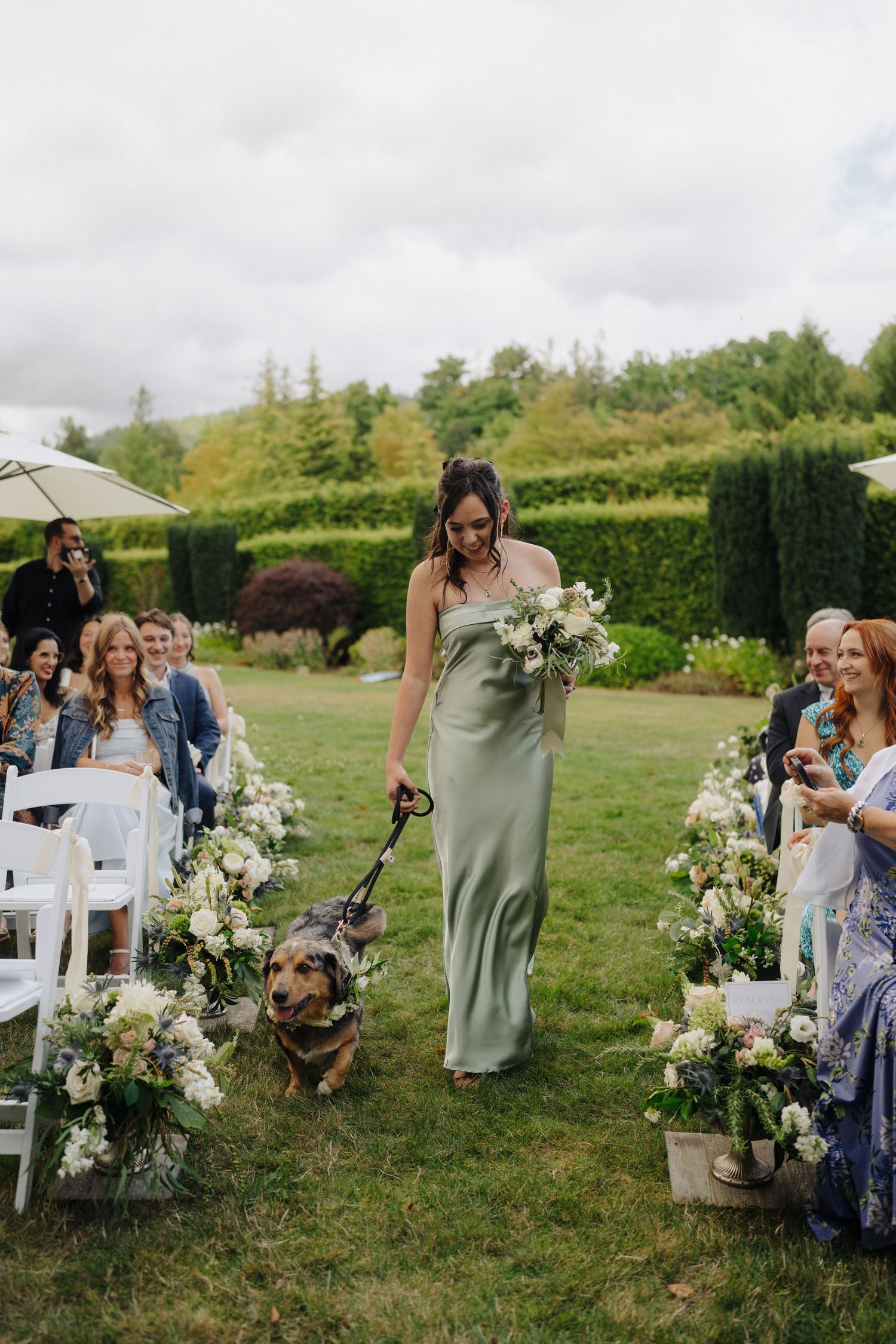 Outdoor wedding ceremony setup with white chairs and floral arrangements along the aisle in a garden setting.