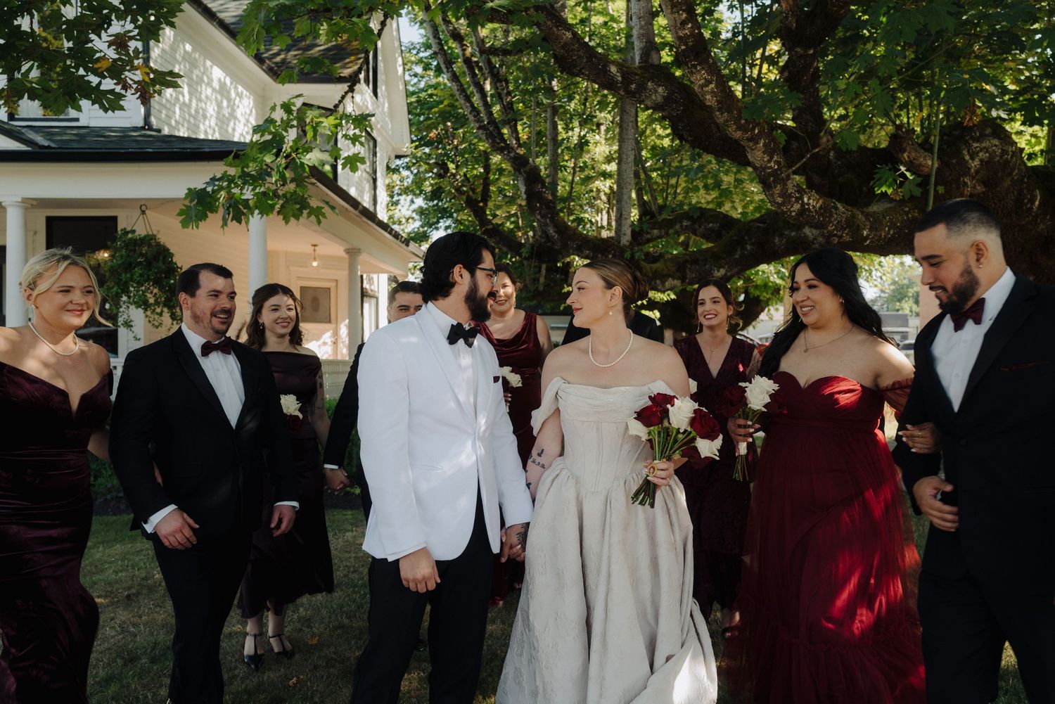 Wedding party gathered outdoors near white house with burgundy and neutral formal attire.
